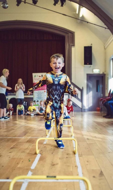 A young boy in a superhero costume is jumping over hurdles in a gym.