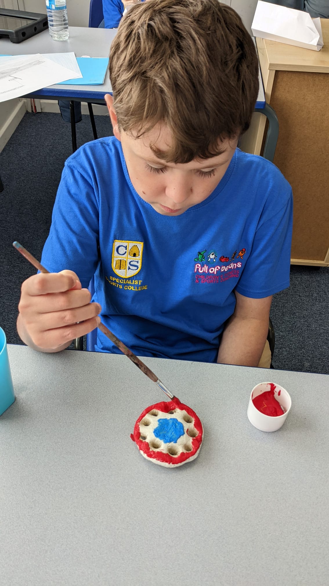 A young boy is sitting at a table painting a red circle with a brush.