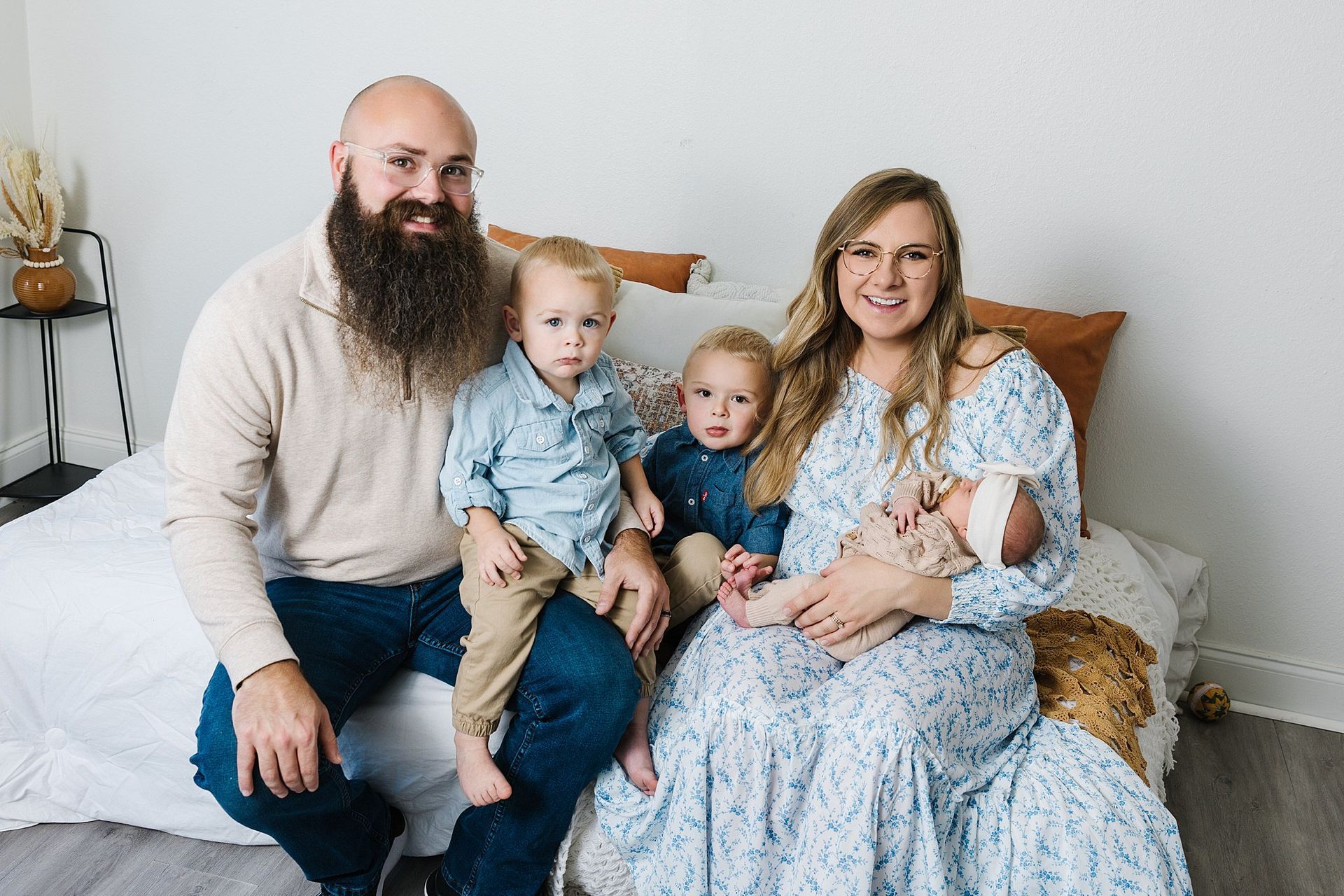 Family of five, posing on a bed: Man with beard, woman holding baby, two boys seated, all smiling.