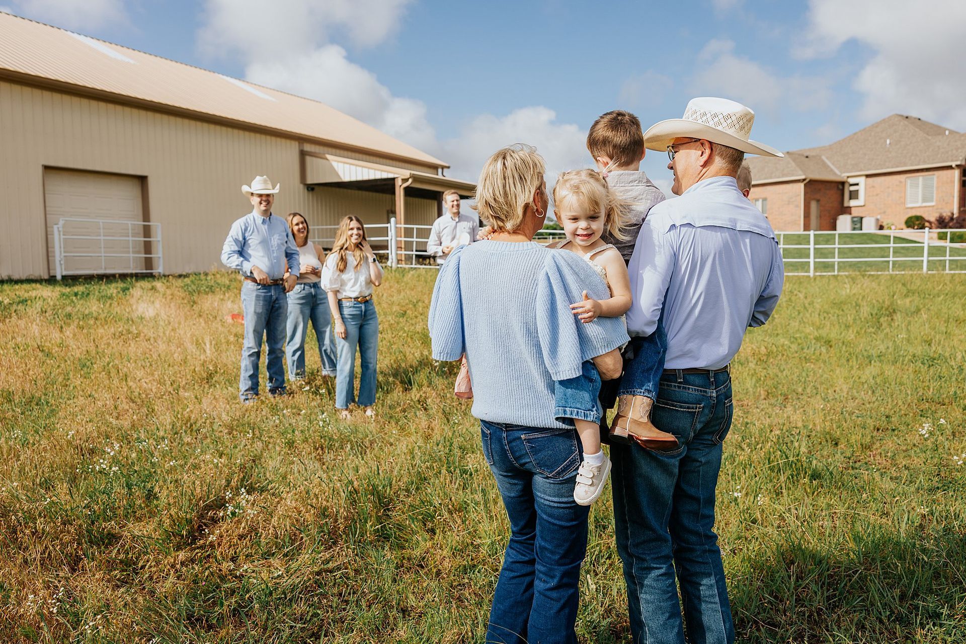 Family posing outdoors in front of a barn and houses. Wearing jeans and cowboy hats, two adults hold children.
