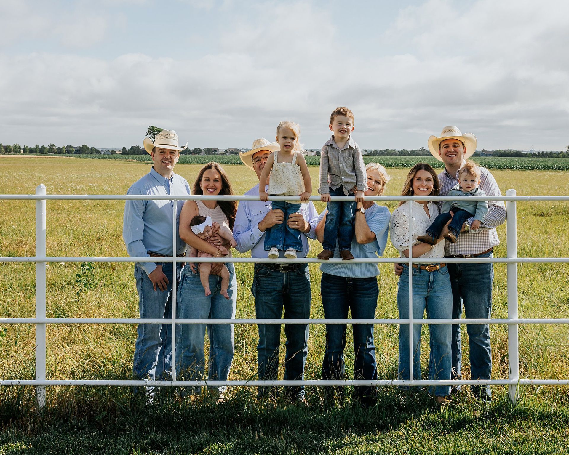 Family posing behind a white fence in a field. Children are held by adults, all wearing cowboy hats.