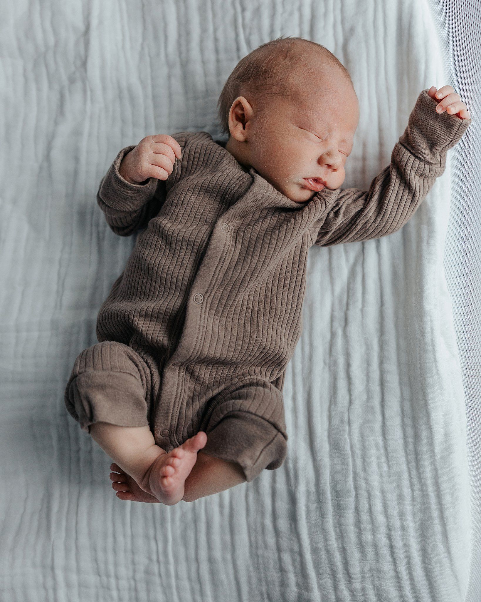 Newborn baby in a brown knit outfit asleep on a white textured blanket.