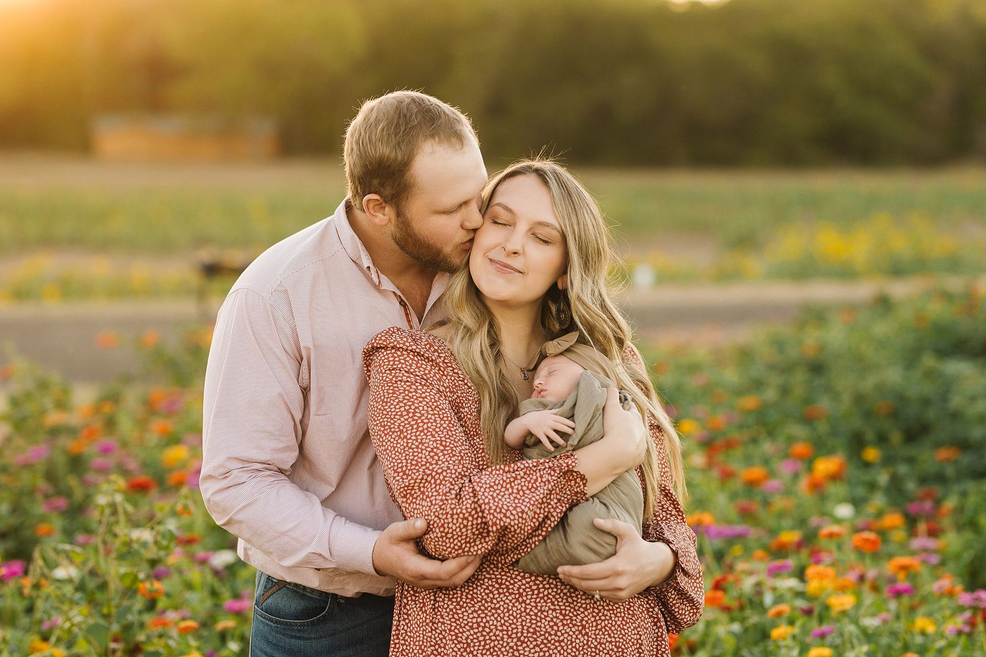 Couple kissing, holding newborn baby in a field of colorful flowers at sunset.