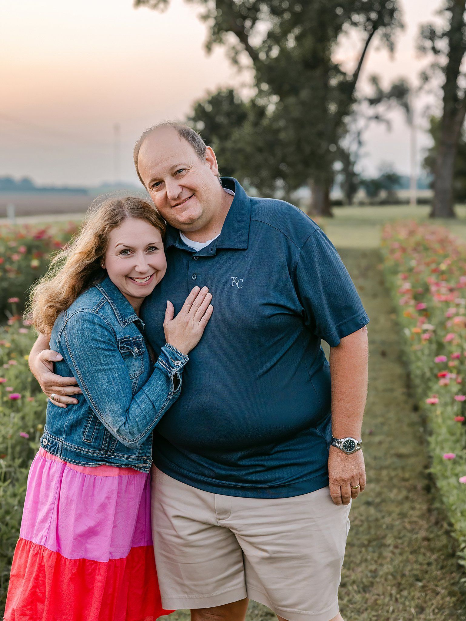 Couple hugging in a flower field; woman in denim jacket and pink dress, man in blue shirt and khakis.