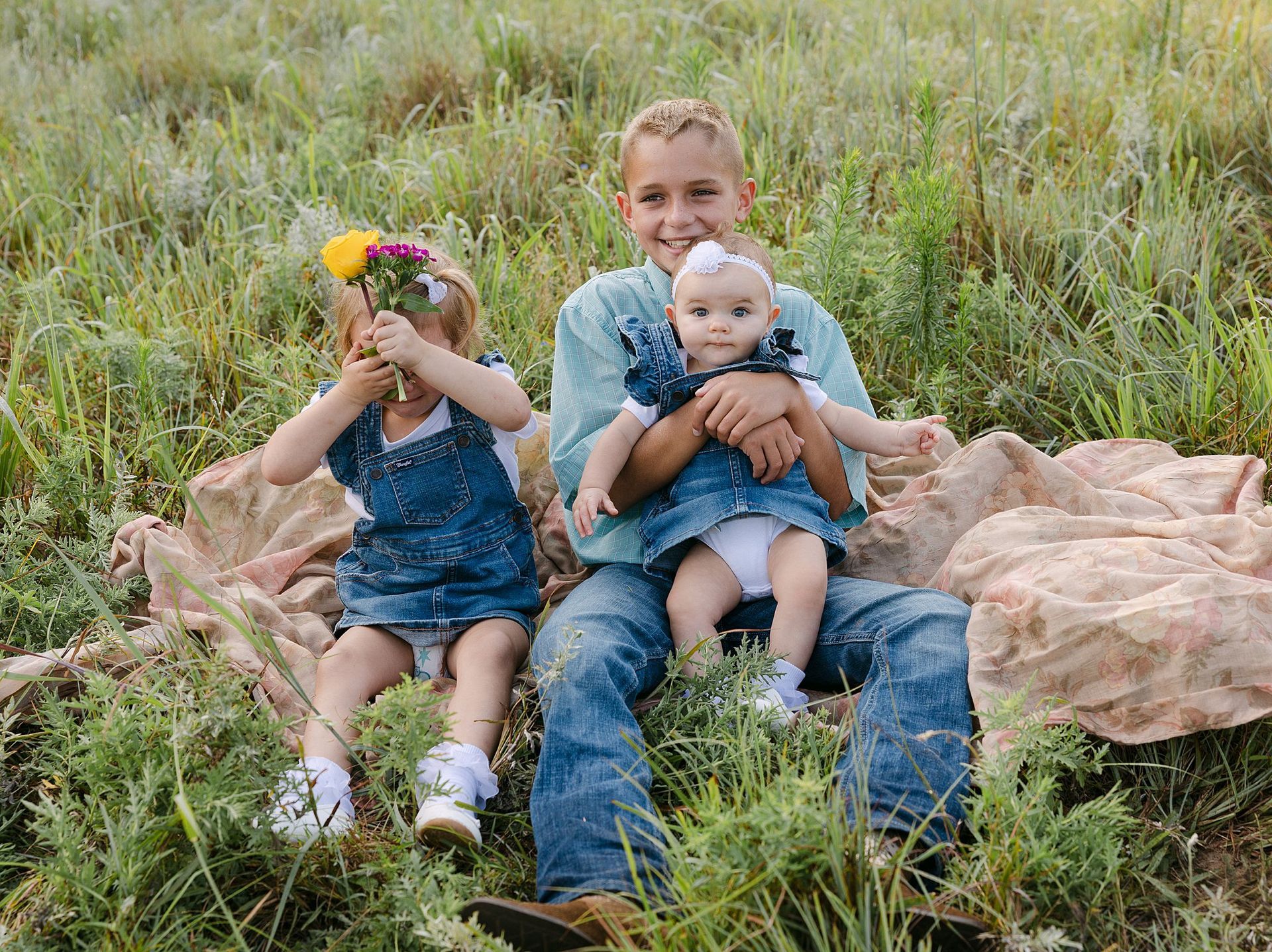Three children in a grassy field: A boy holds a baby, while a girl hides behind flowers.