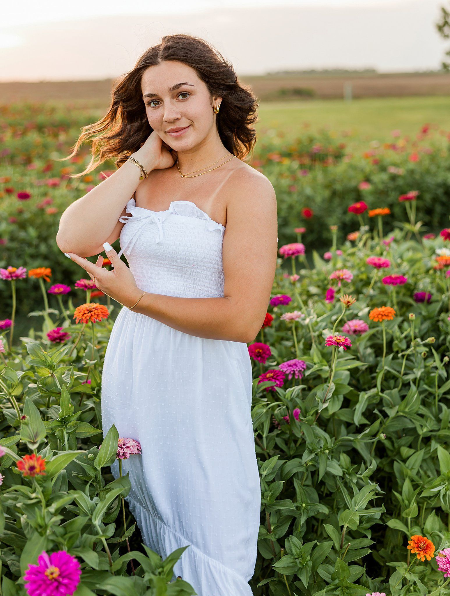 Woman in white dress in a field of colorful flowers, smiling, hand on hair.