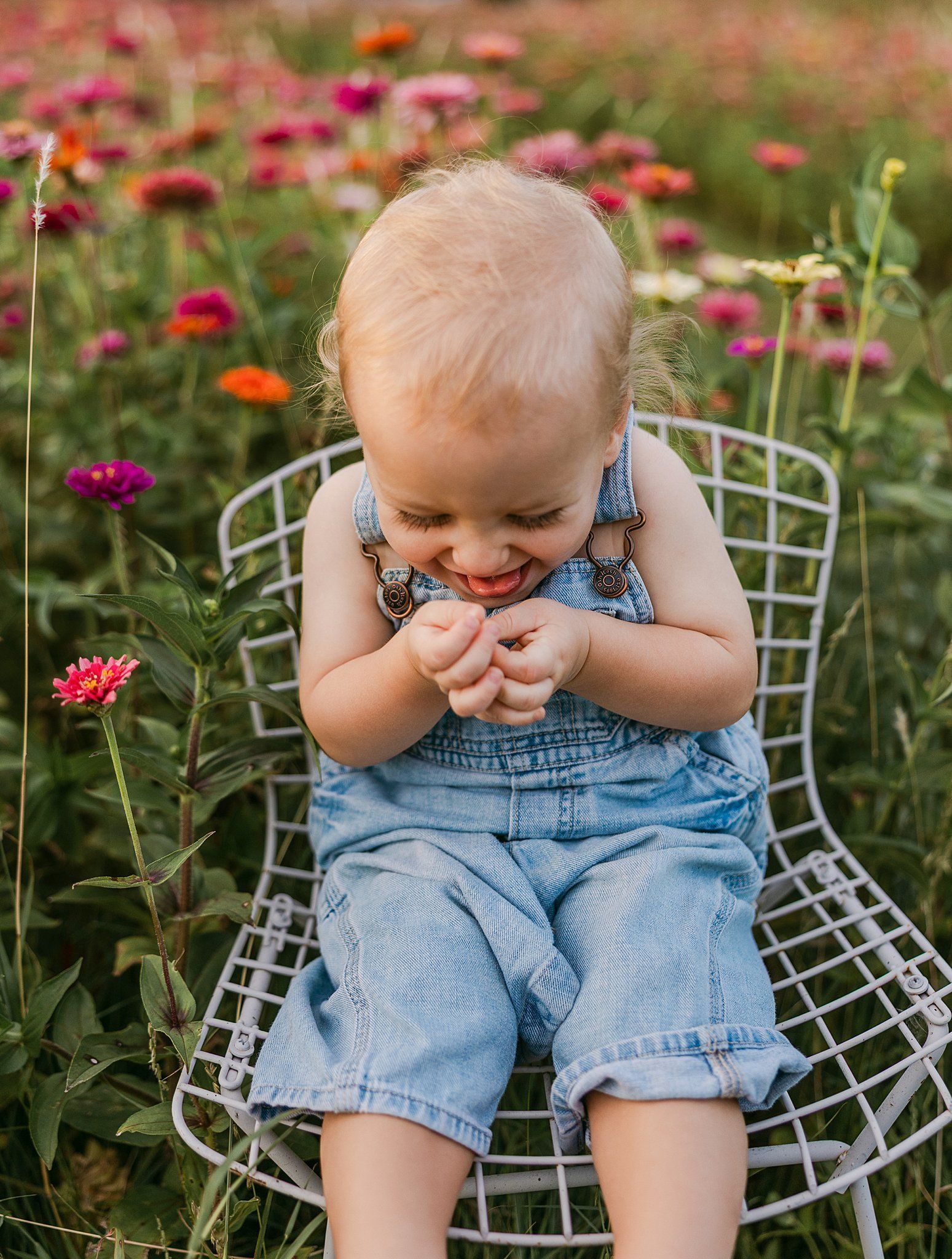 A smiling toddler in blue overalls sits on a white chair in a field of colorful flowers.