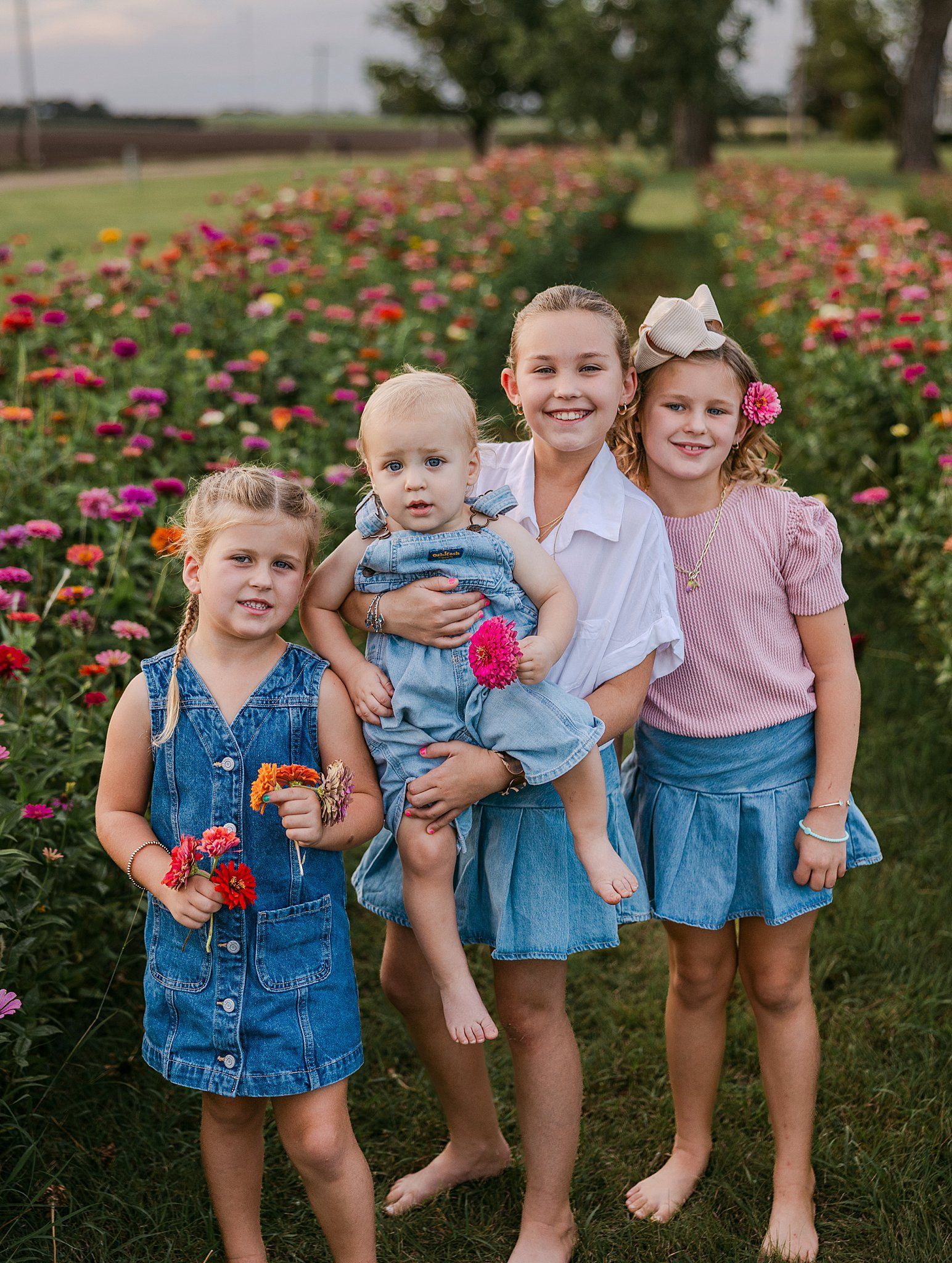 Four young girls smiling in a colorful flower field, one holding a baby, all wearing denim.