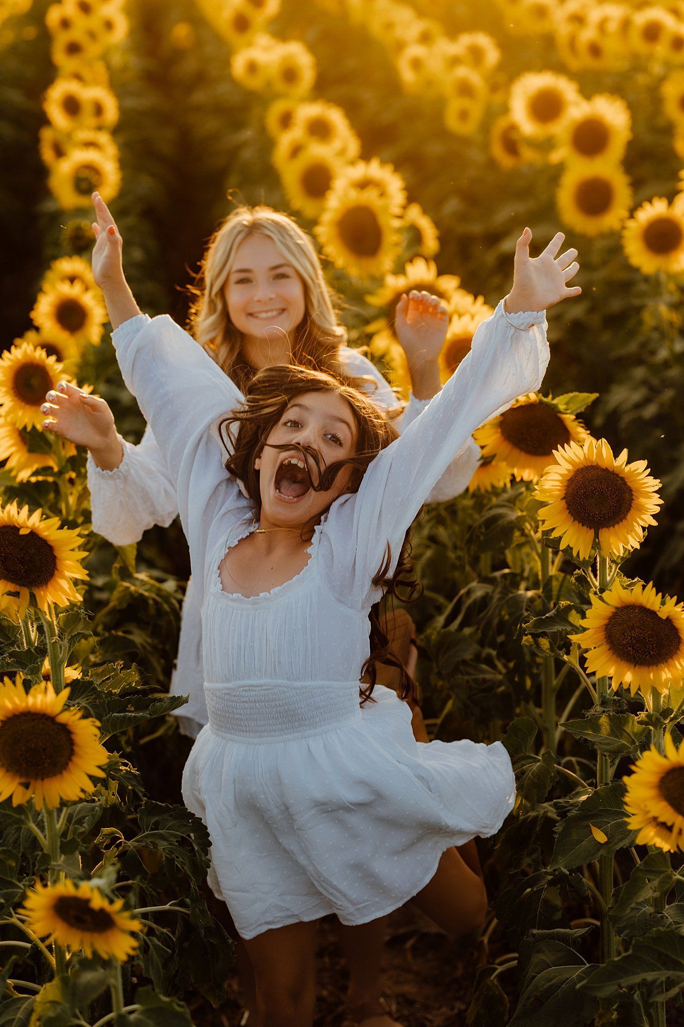 Two girls in white dresses joyfully in a sunflower field, arms raised, sunny lighting.