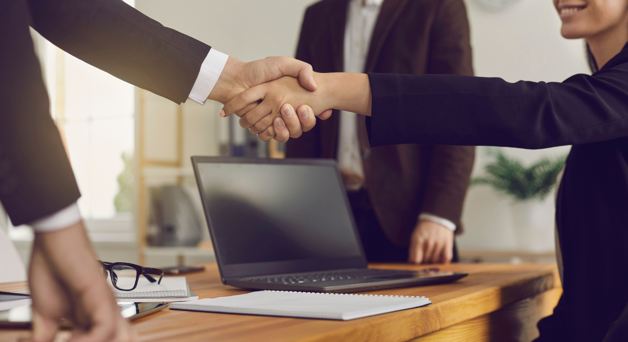 Two people shaking hands across a desk with a laptop in a meeting room