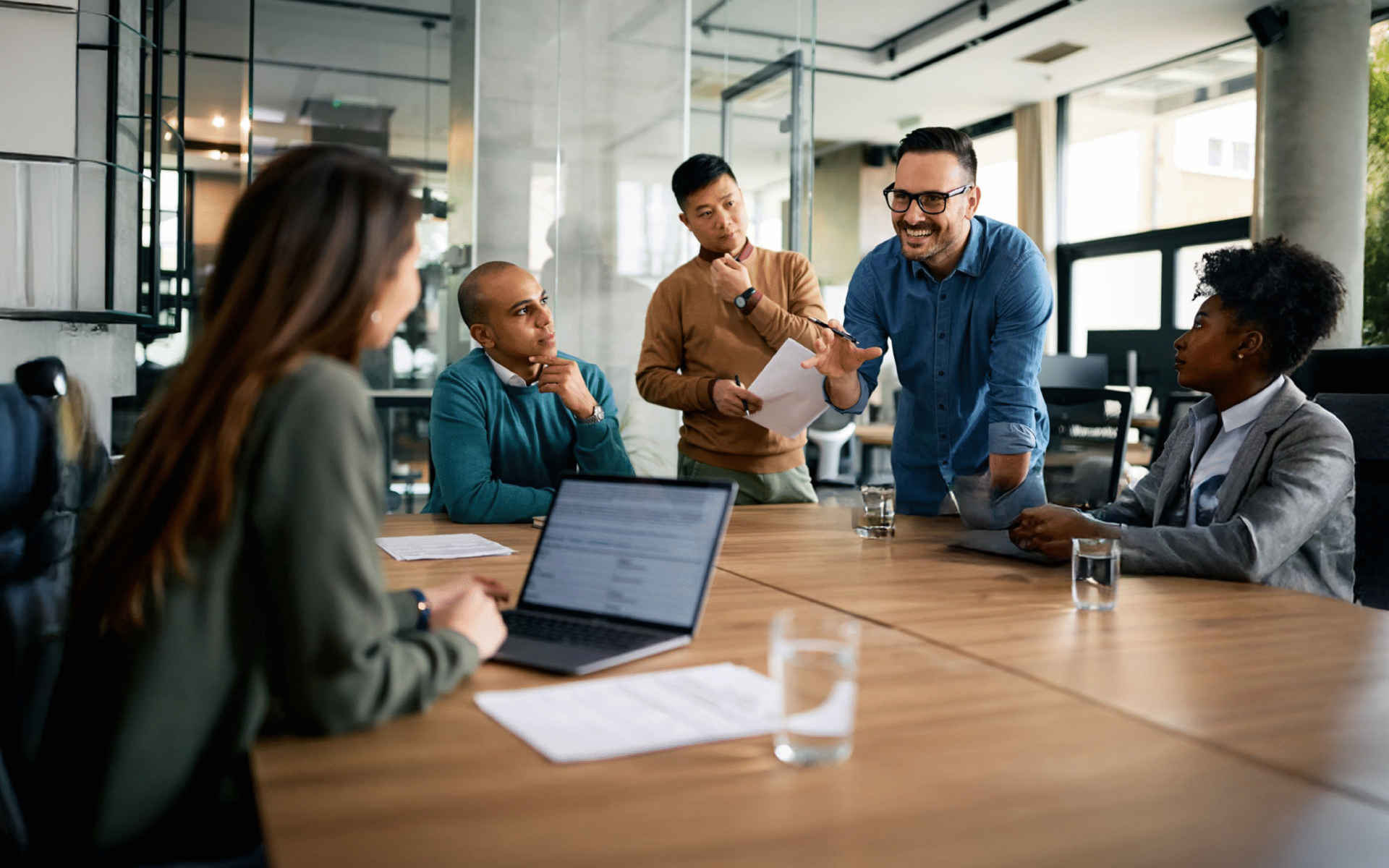 Team meeting in a bright office, with five people discussing around a wooden conference table.