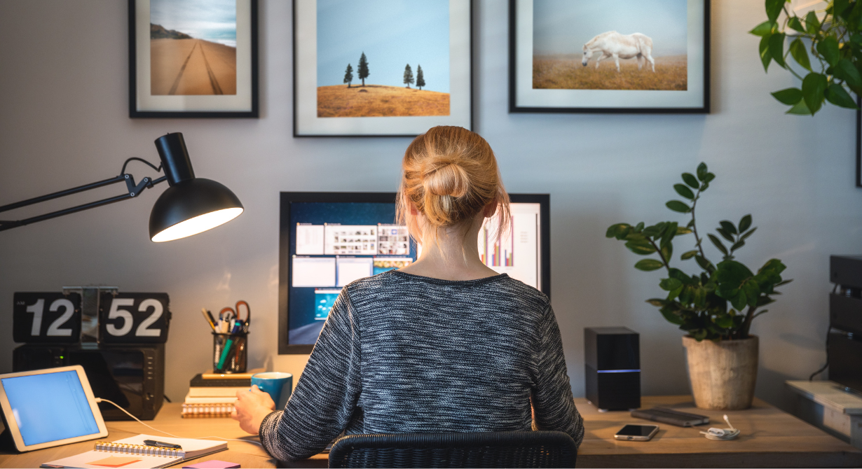 Person working at a desk with dual monitors, desk lamp, clock, and potted plants in a home office
