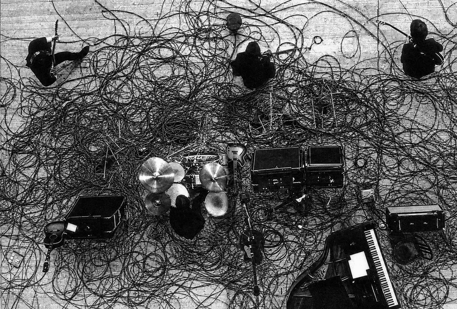Black-and-white scene of tangled wires and scattered objects on a table, with several small figures around it