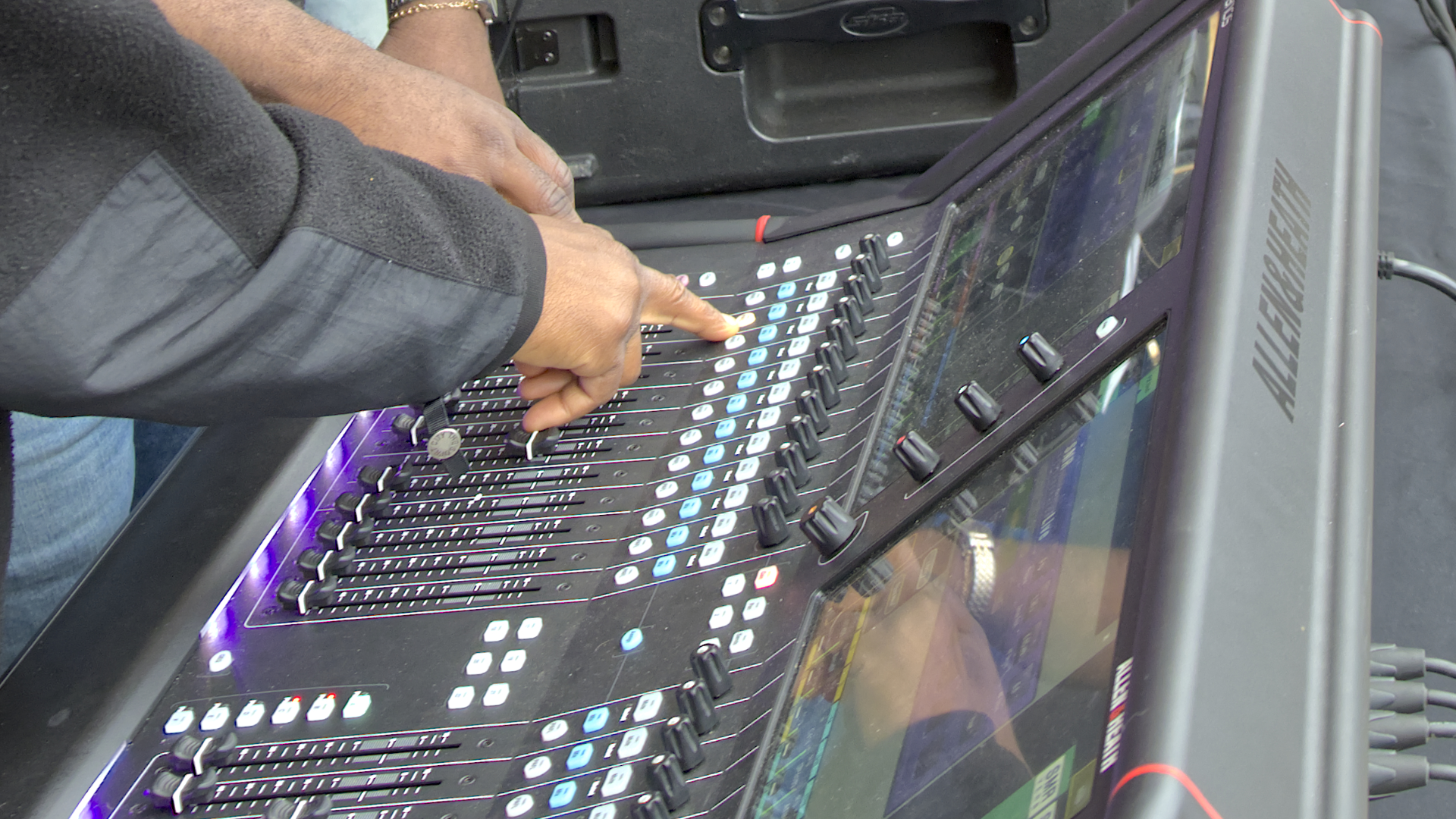 Hands adjusting faders and buttons on a large, modern sound mixing console, likely at an outdoor event.