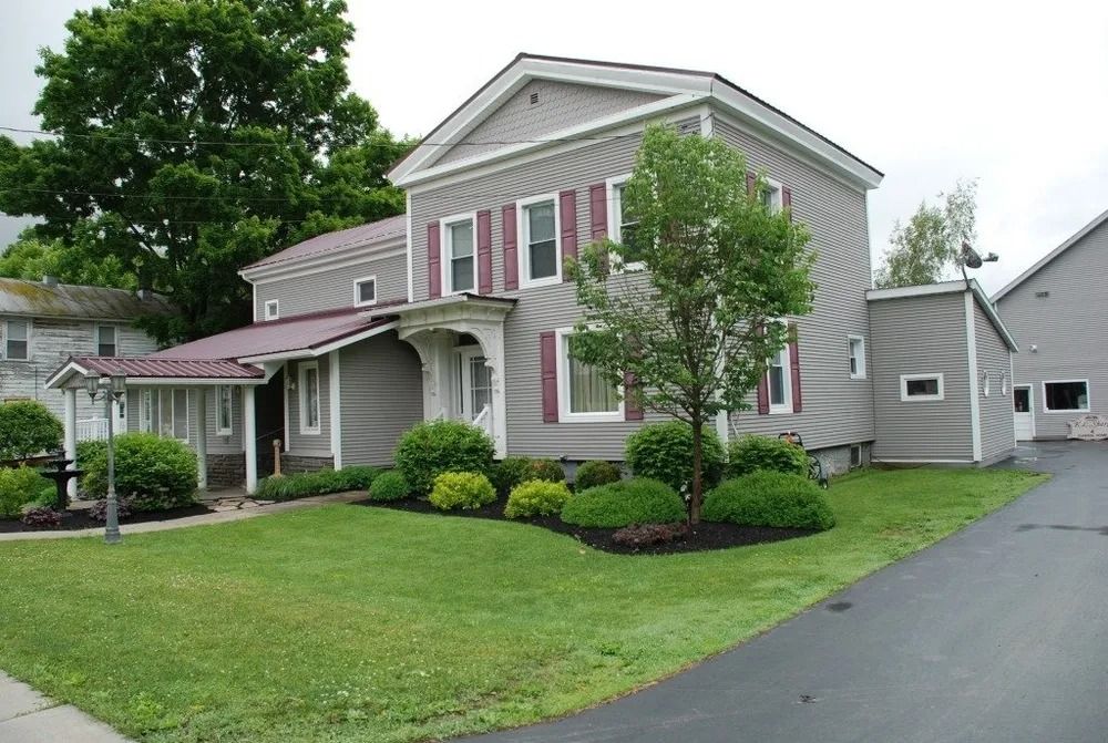 Two-story gray house with red shutters and metal roof. Front yard with green grass and small trees.