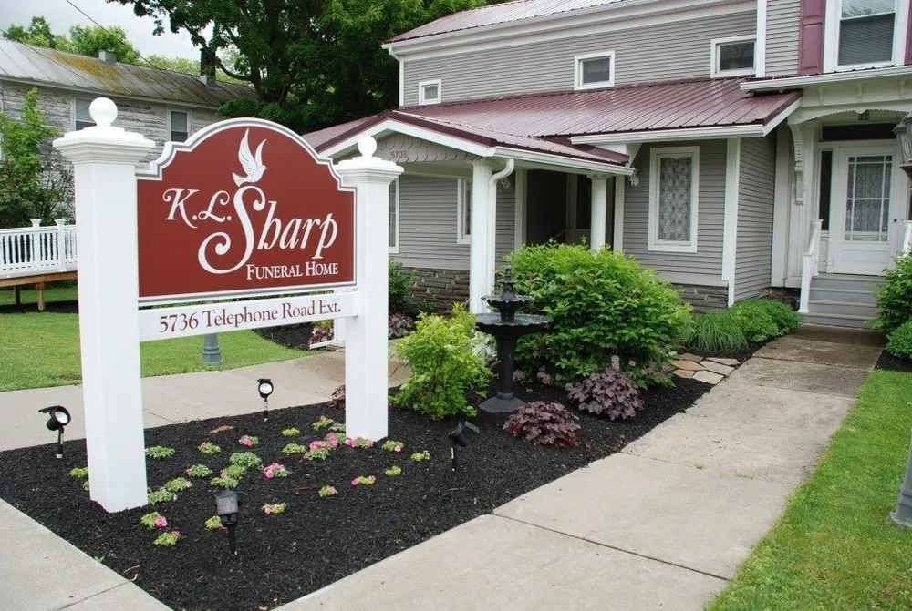 K.L. Sharp Funeral Home sign with address, in front of a gray building with a red roof. Landscaping in front.