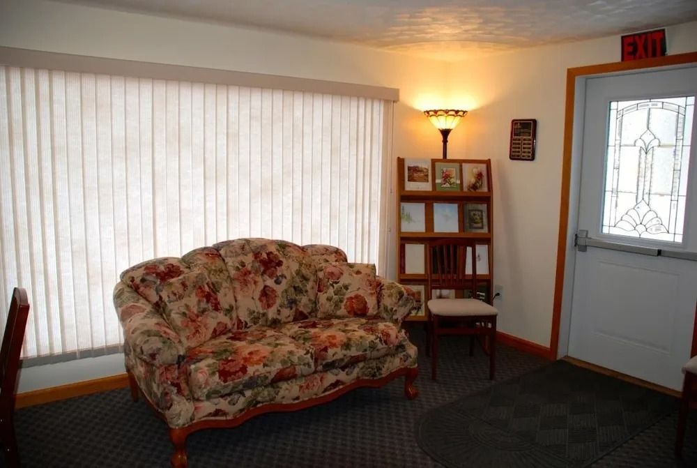 Floral couch by window, small bookcase, floor lamp, exit door. Interior with white blinds, dark carpet.