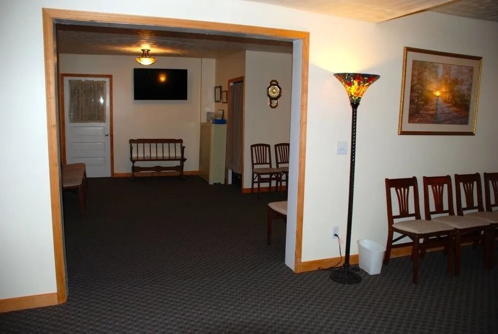 Hallway with dark carpet, wooden chairs, and a floor lamp. A doorway leads to another room.