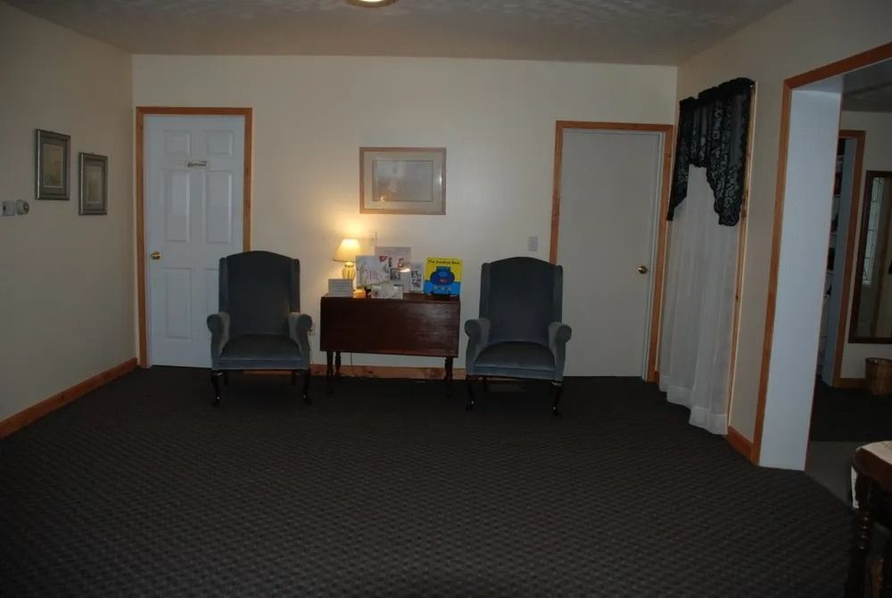 Two upholstered chairs face a small table. Two doors, art, and a curtain are visible in a beige room with dark carpet.