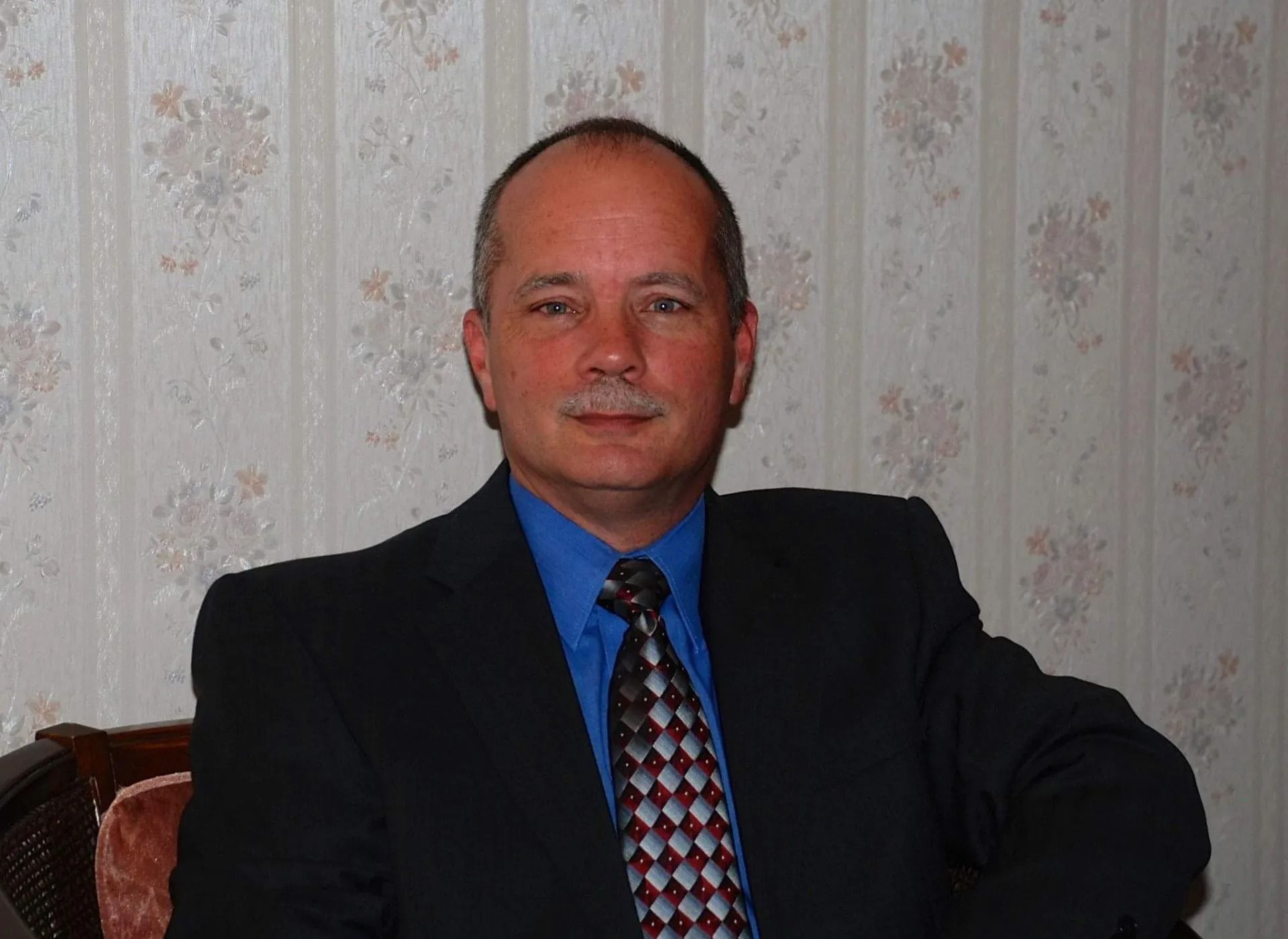 Man in suit, seated, smiling. Floral wallpaper background.