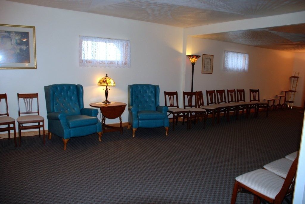 A funeral home waiting room with blue armchairs, rows of wooden chairs, and a floor lamp.