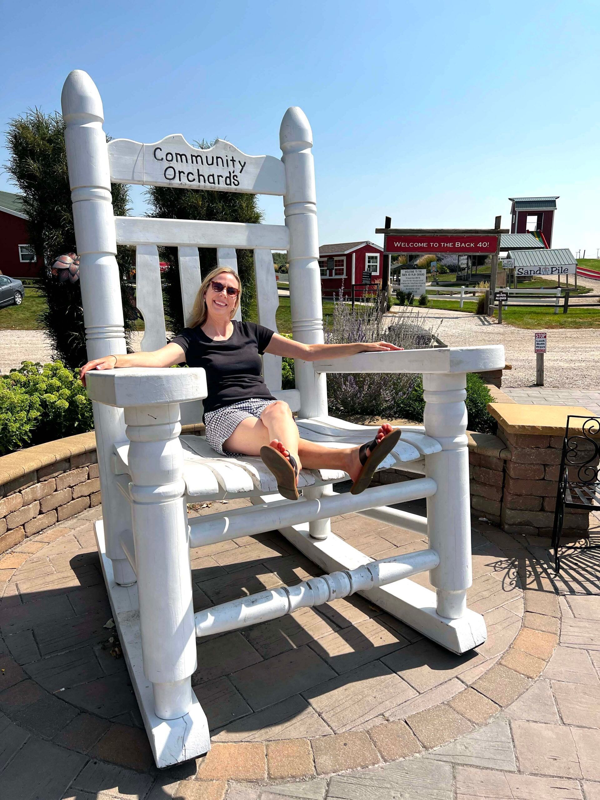 woman in very oversized white rocking chair outdoors with a red barn and playground in the background
