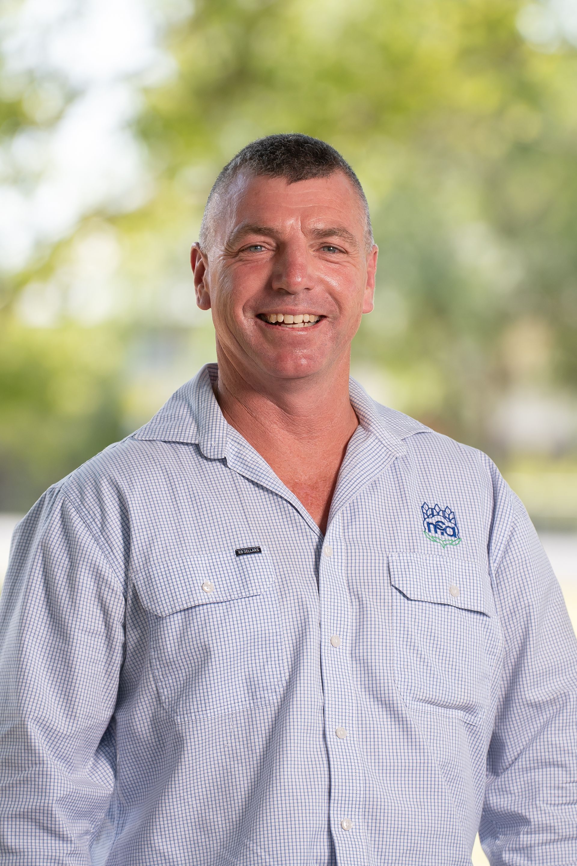 Man smiling, wearing a light blue button-down shirt with logo on chest, outdoors.