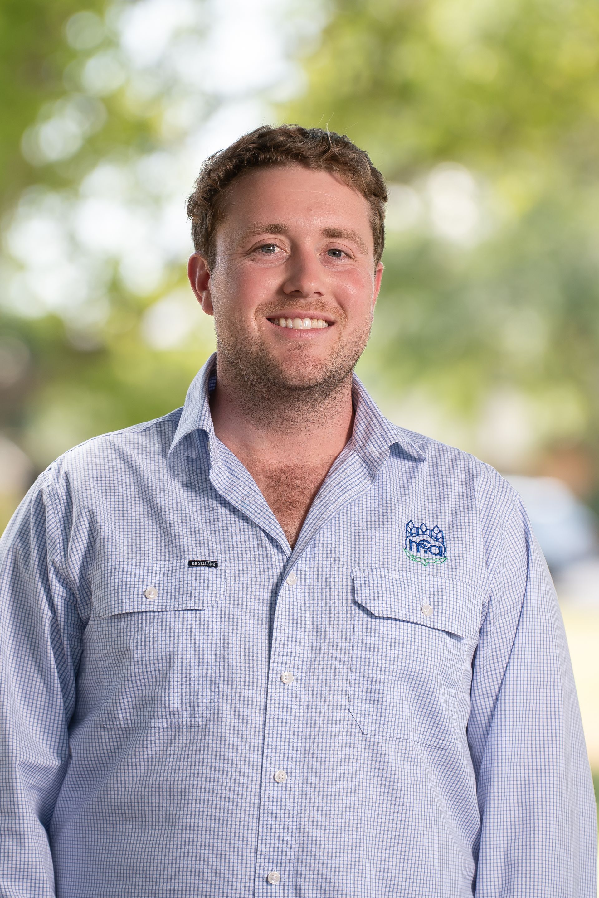 Man wearing a blue striped button-up shirt smiles in front of a blurred green background.