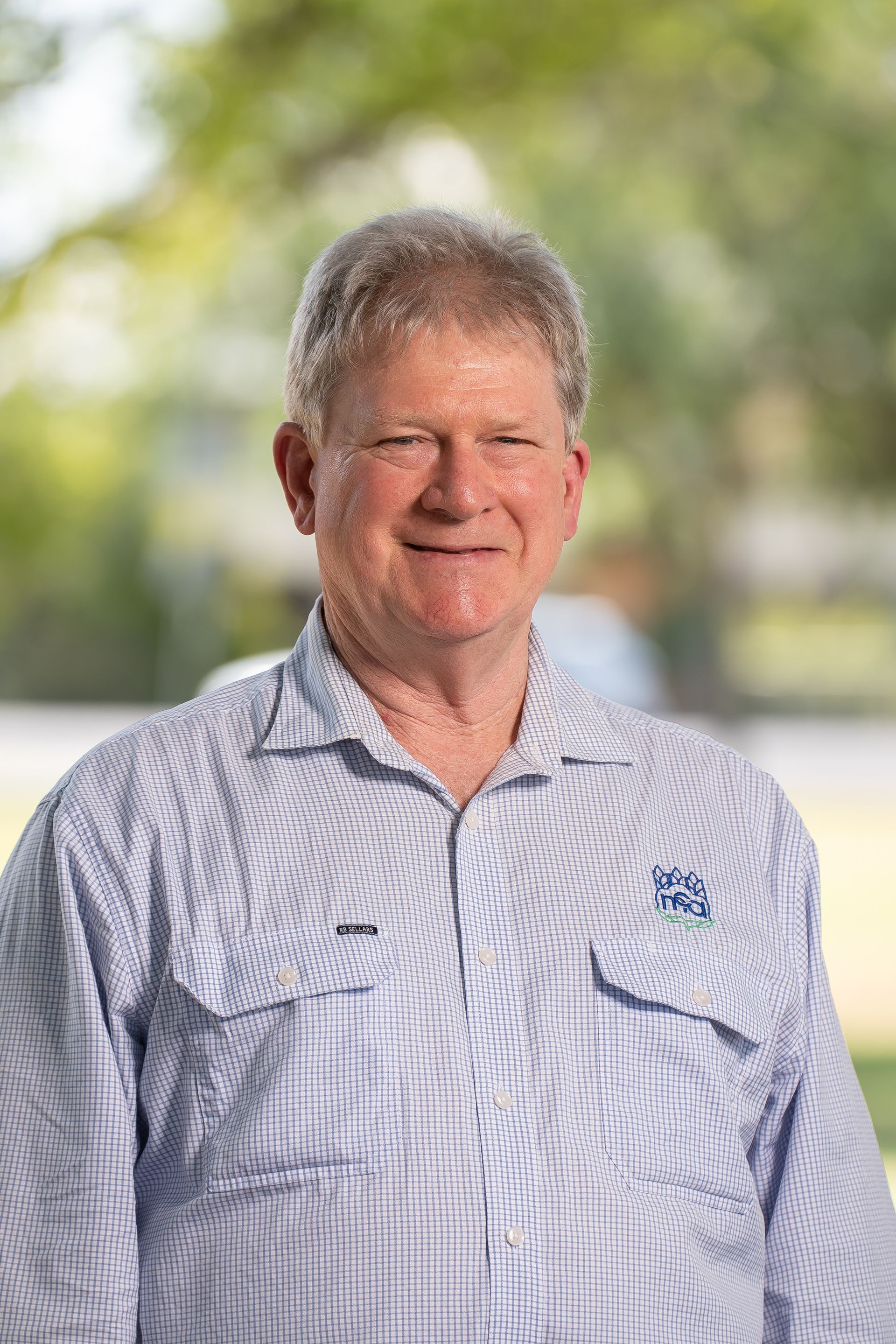 Man in light blue shirt smiling outdoors, blurred background.