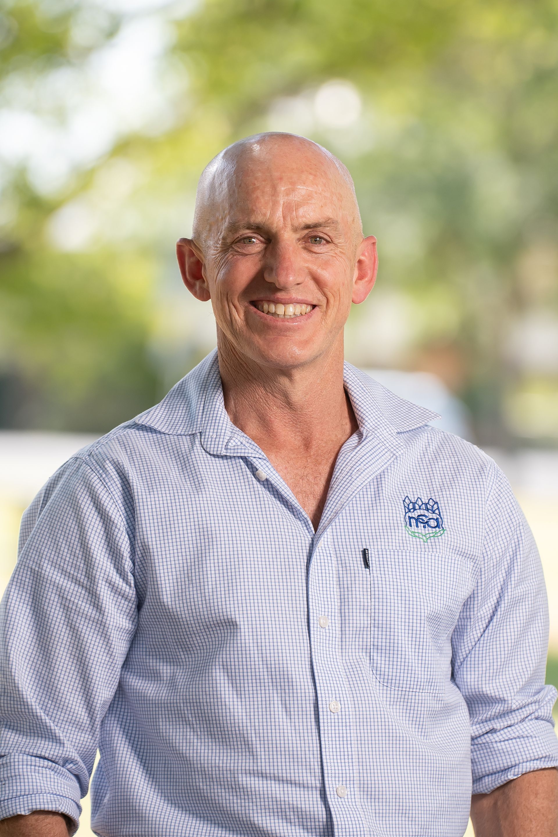 Bald man smiling, wearing light blue button-down shirt outdoors.