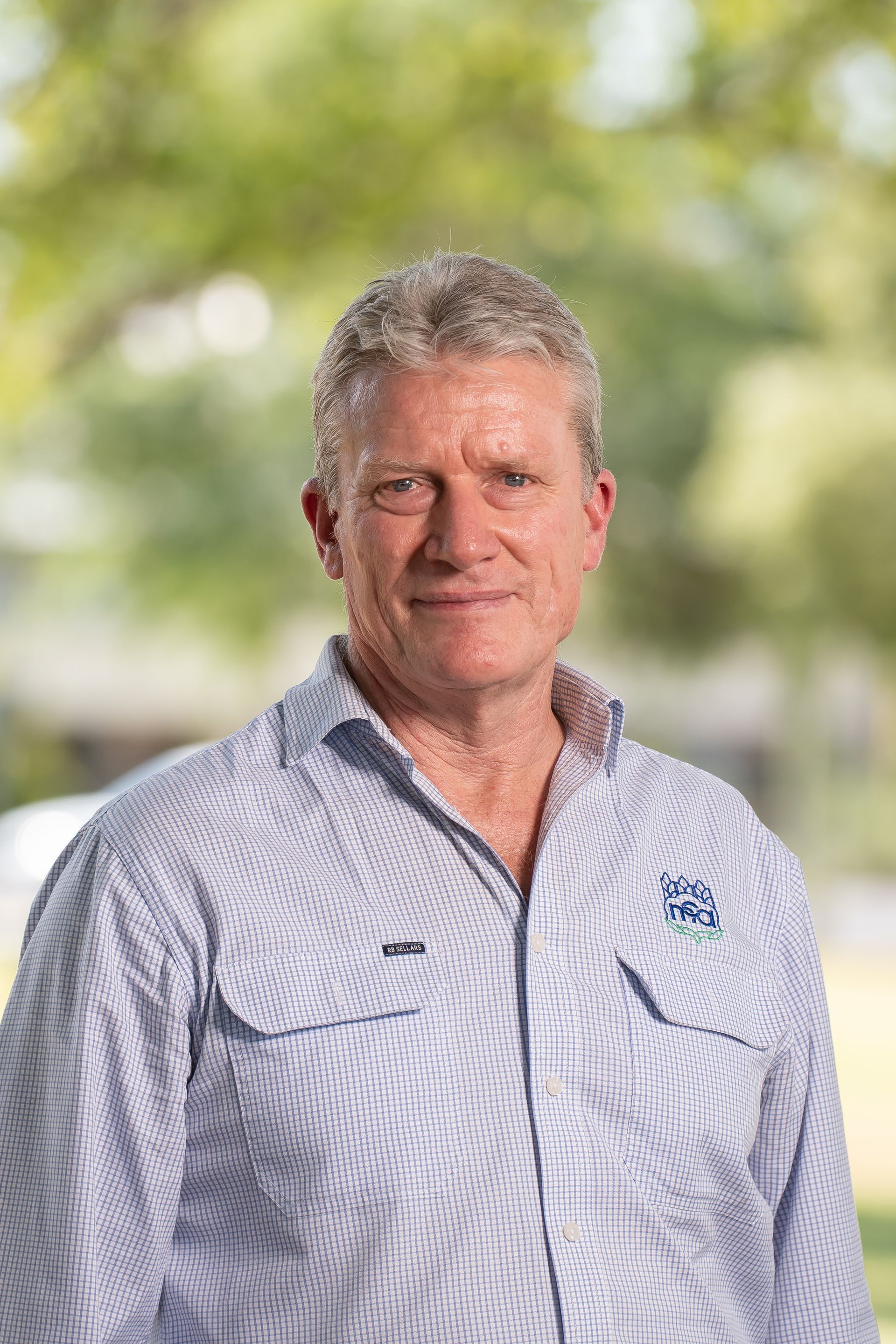 Man in a light blue collared shirt smiling, outdoors with a blurred background of trees.
