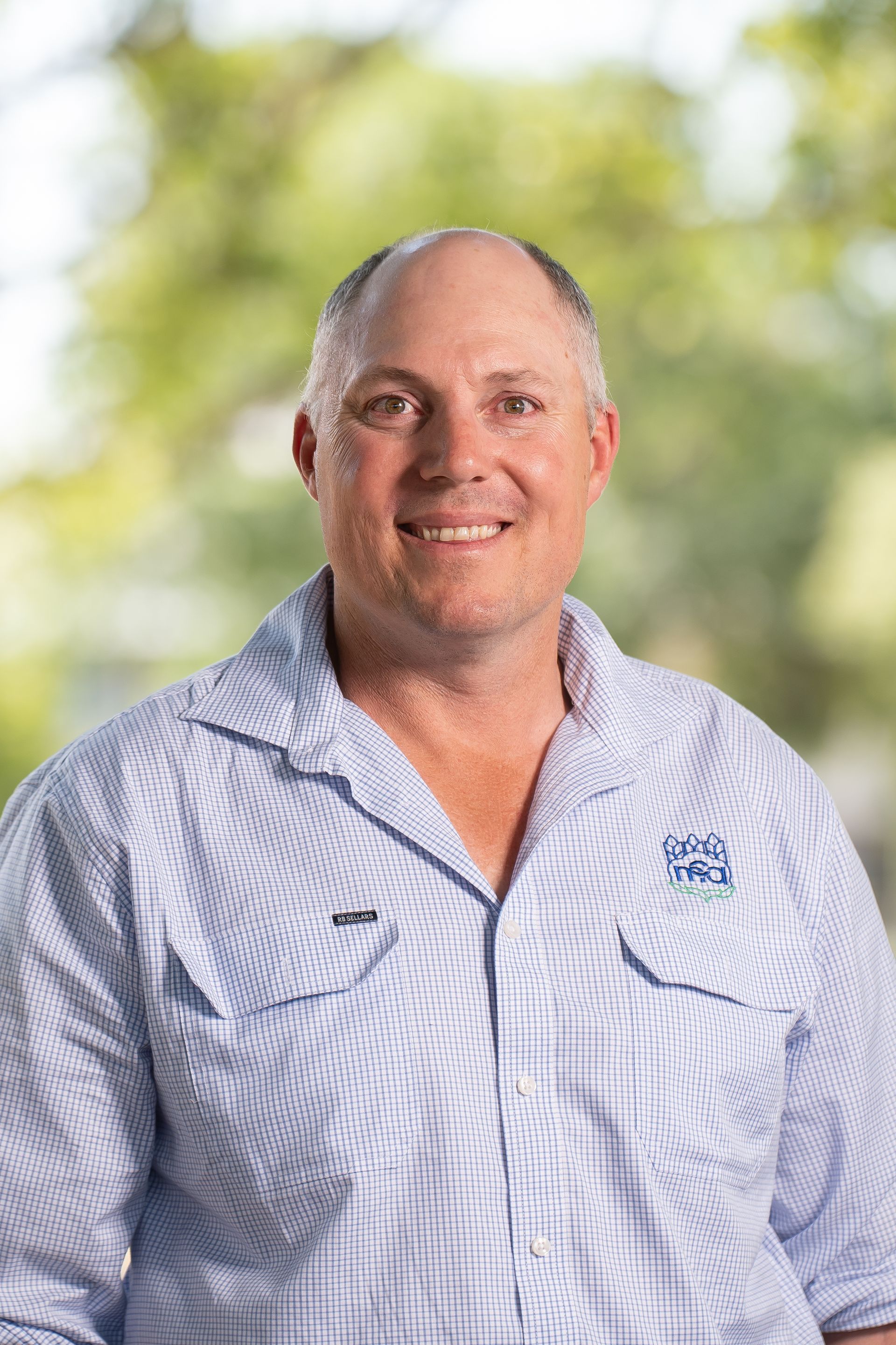 Man with fair skin wearing a blue button-down shirt smiles in front of a blurred green background.