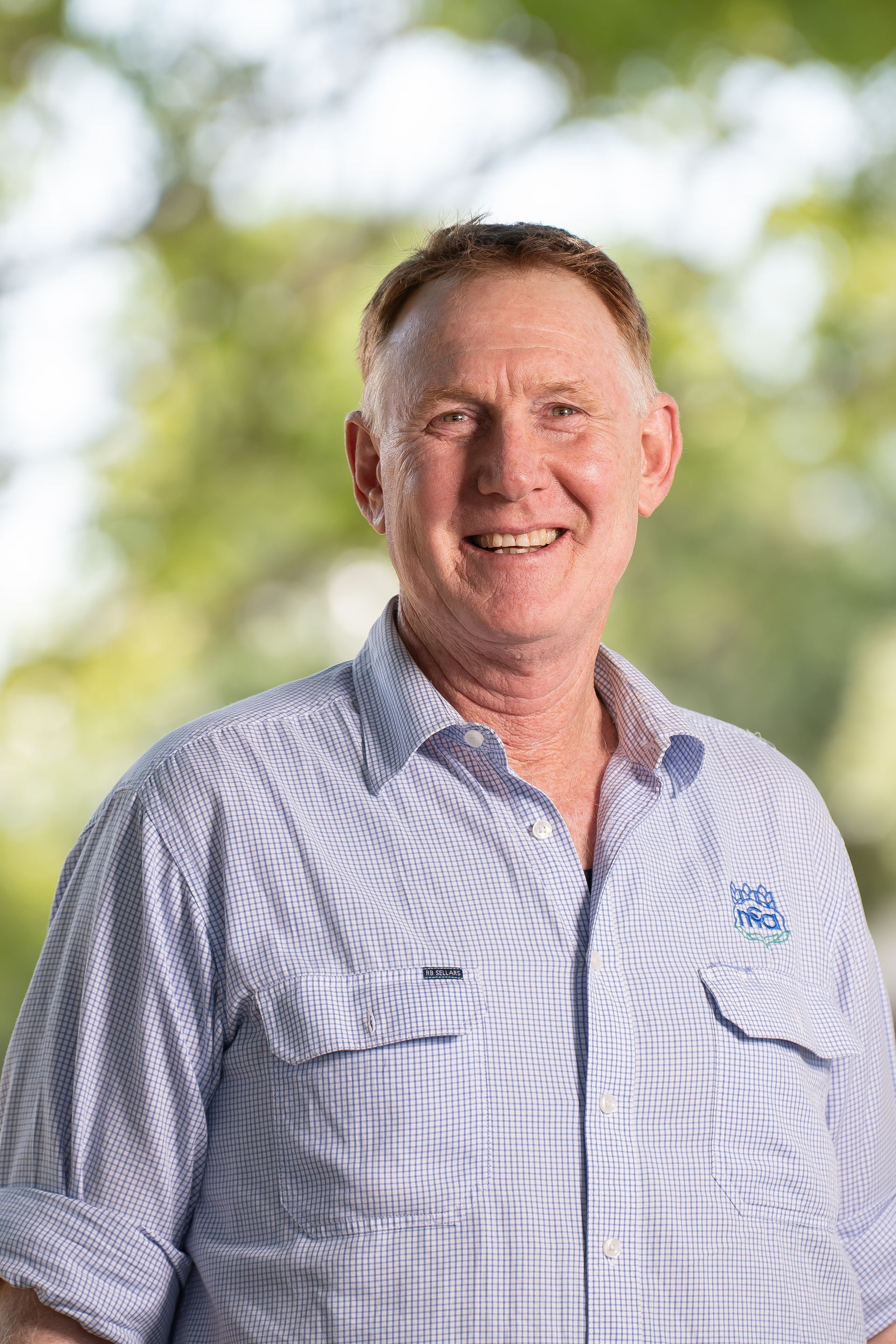 Man in a blue and white collared shirt smiles, set against a blurred green backdrop.