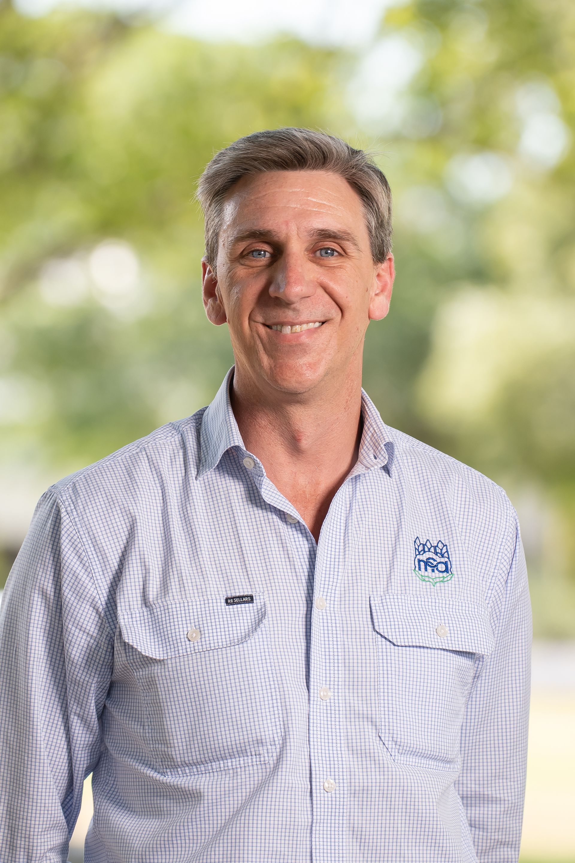 Man smiling in a light blue patterned shirt outdoors with a blurred green background.