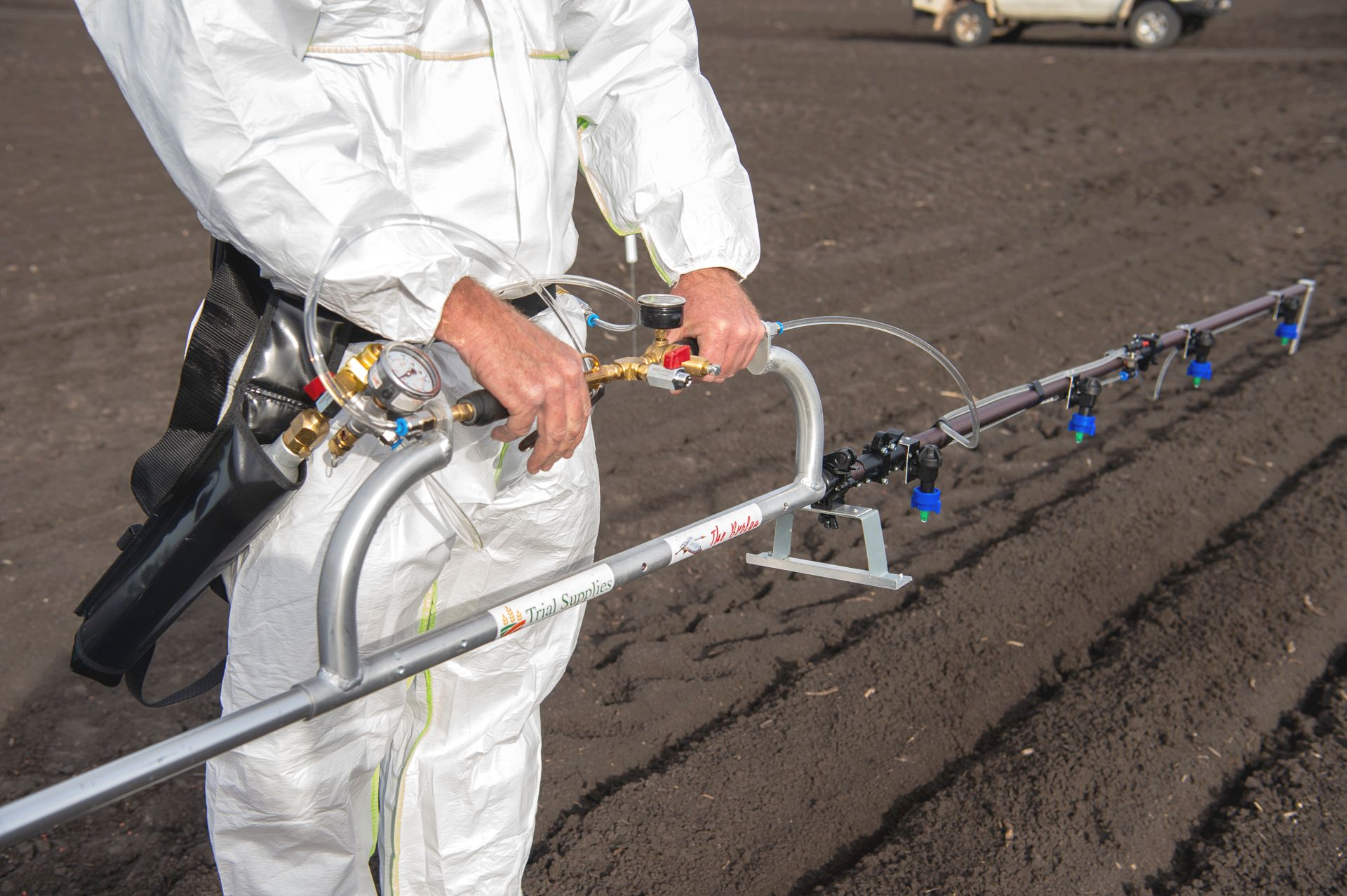 Person in protective suit sprays crops with a hand-held sprayer in a field.