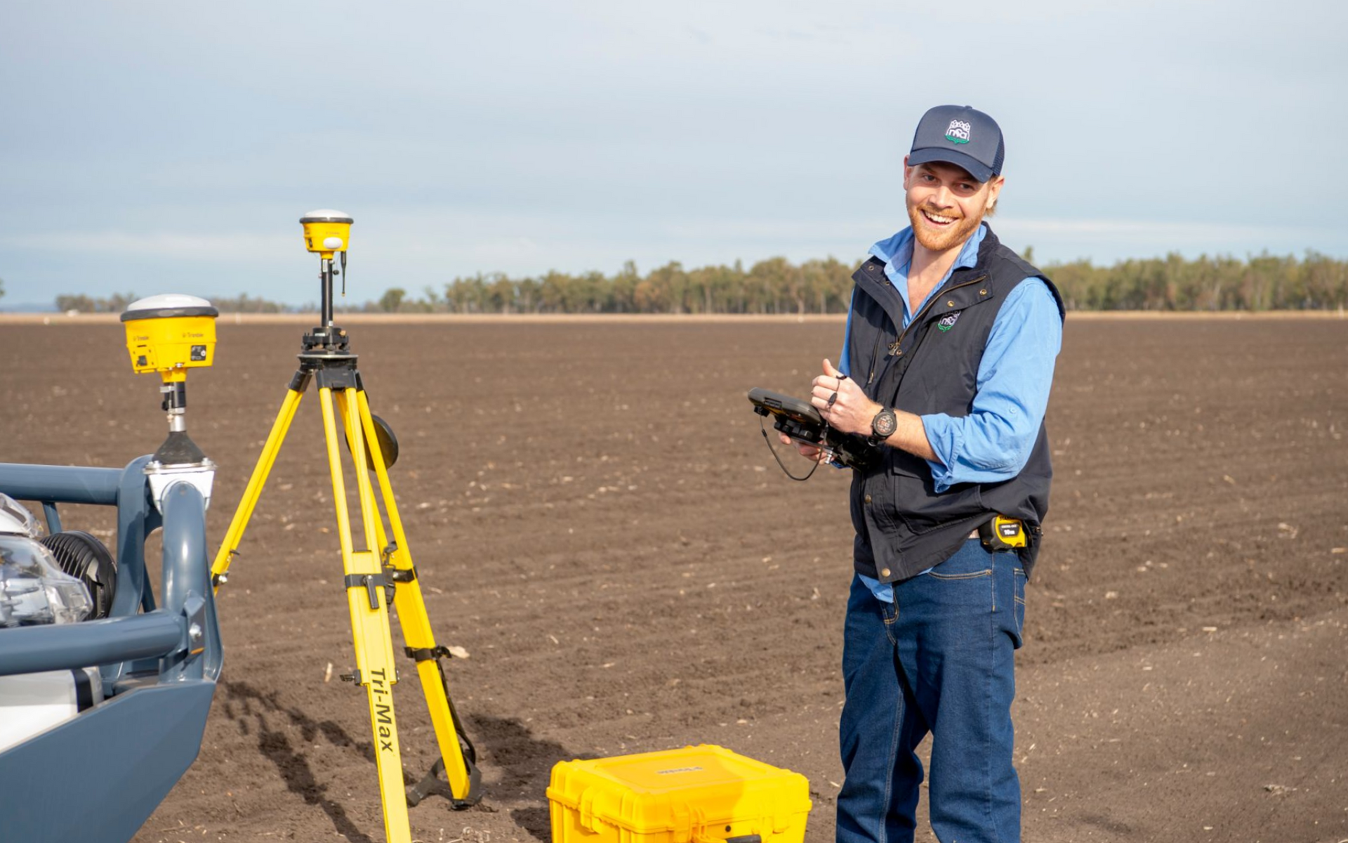 Man in a field with surveying equipment, smiling and holding a tablet. Sunny day.