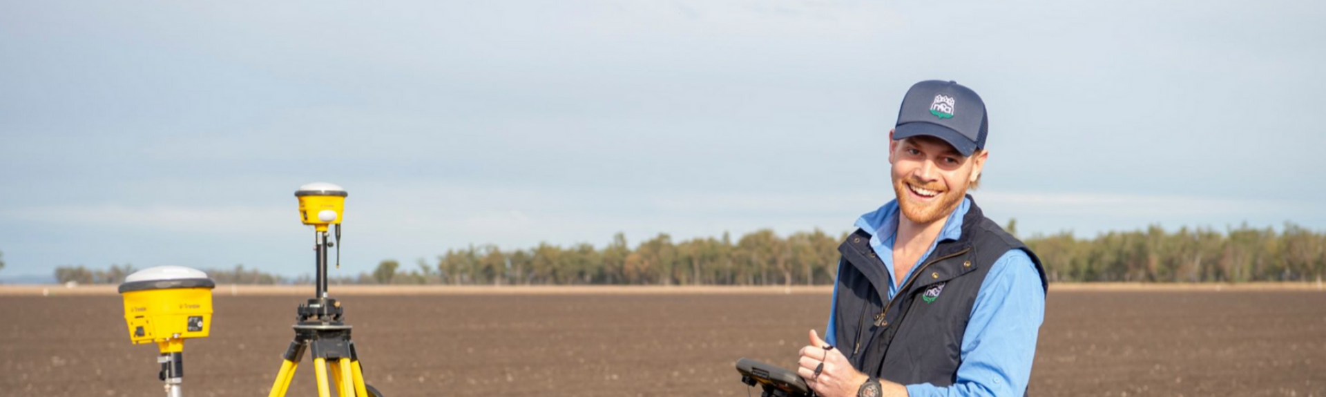 Man in field with surveying equipment, smiling. Brown field, grey sky.