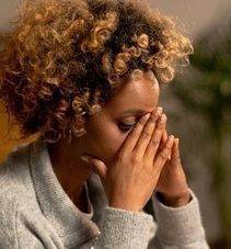 Woman with dark skin sits on a gray couch, looking sad, hand on her head.