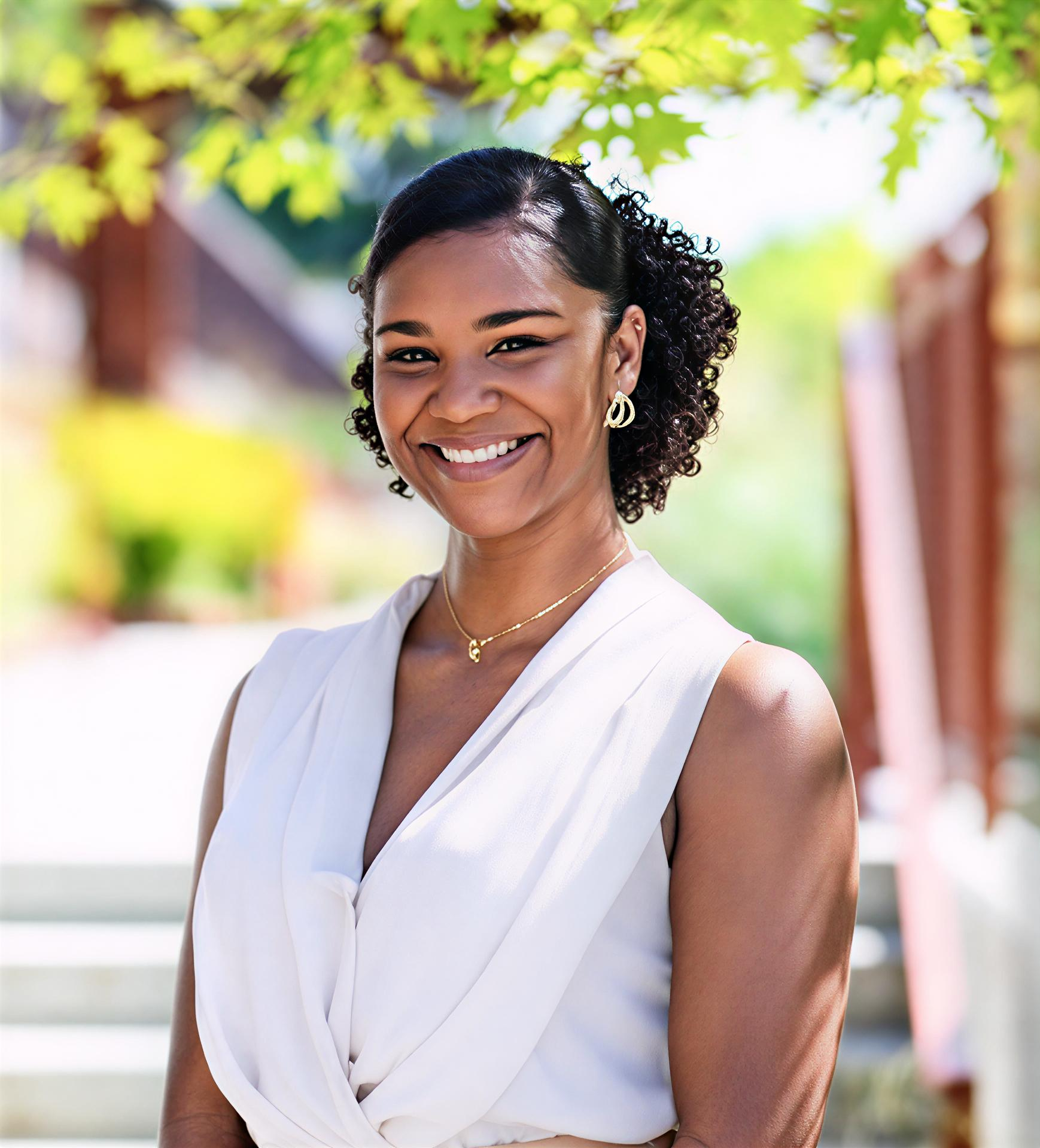 Woman smiling, outdoors, wearing a sleeveless cream top, small gold necklace and earrings, with curly hair up.