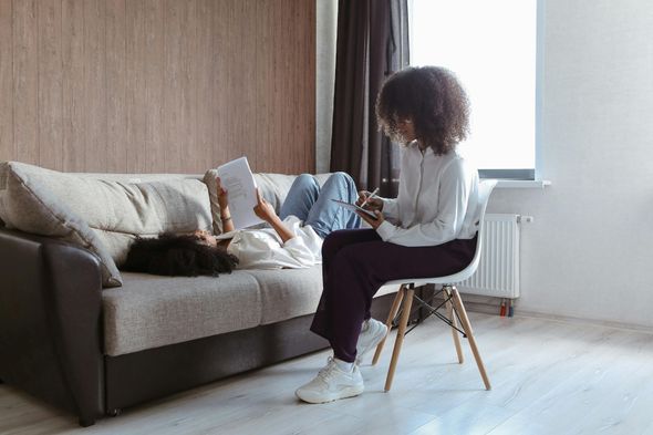 A woman is sitting on a chair in front of a couch reading a book.