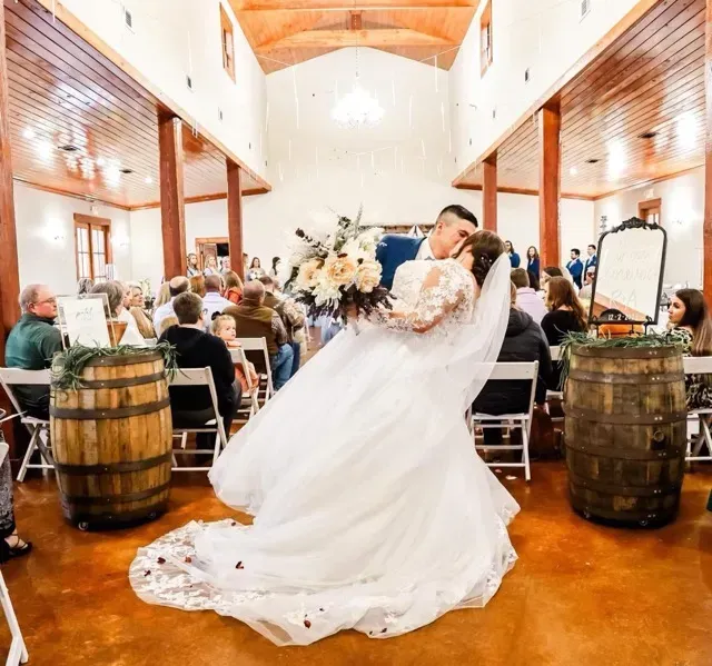 Bride and groom kissing in a rustic wedding hall, surrounded by seated guests and wooden barrels