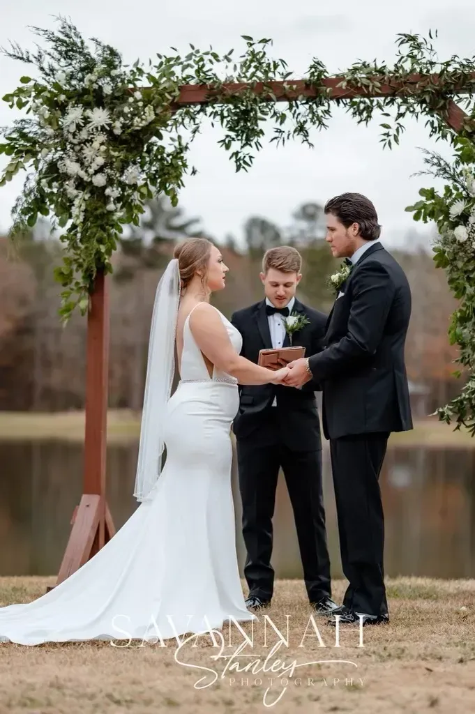 Bride and groom exchanging vows under a wooden arch by a lake, with an officiant nearby.