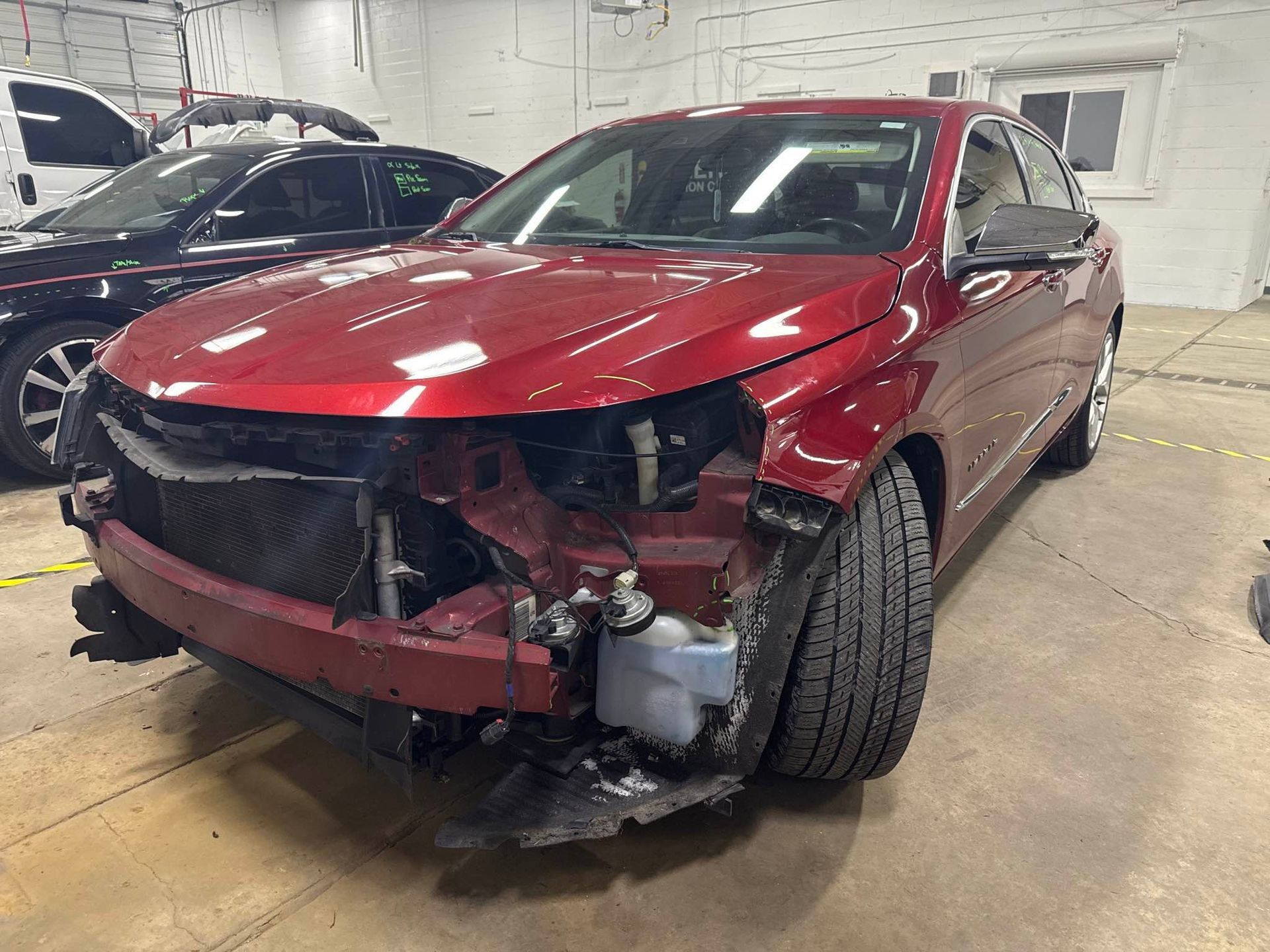 Red damaged sedan in a garage, front-end collision, missing parts on the left side.