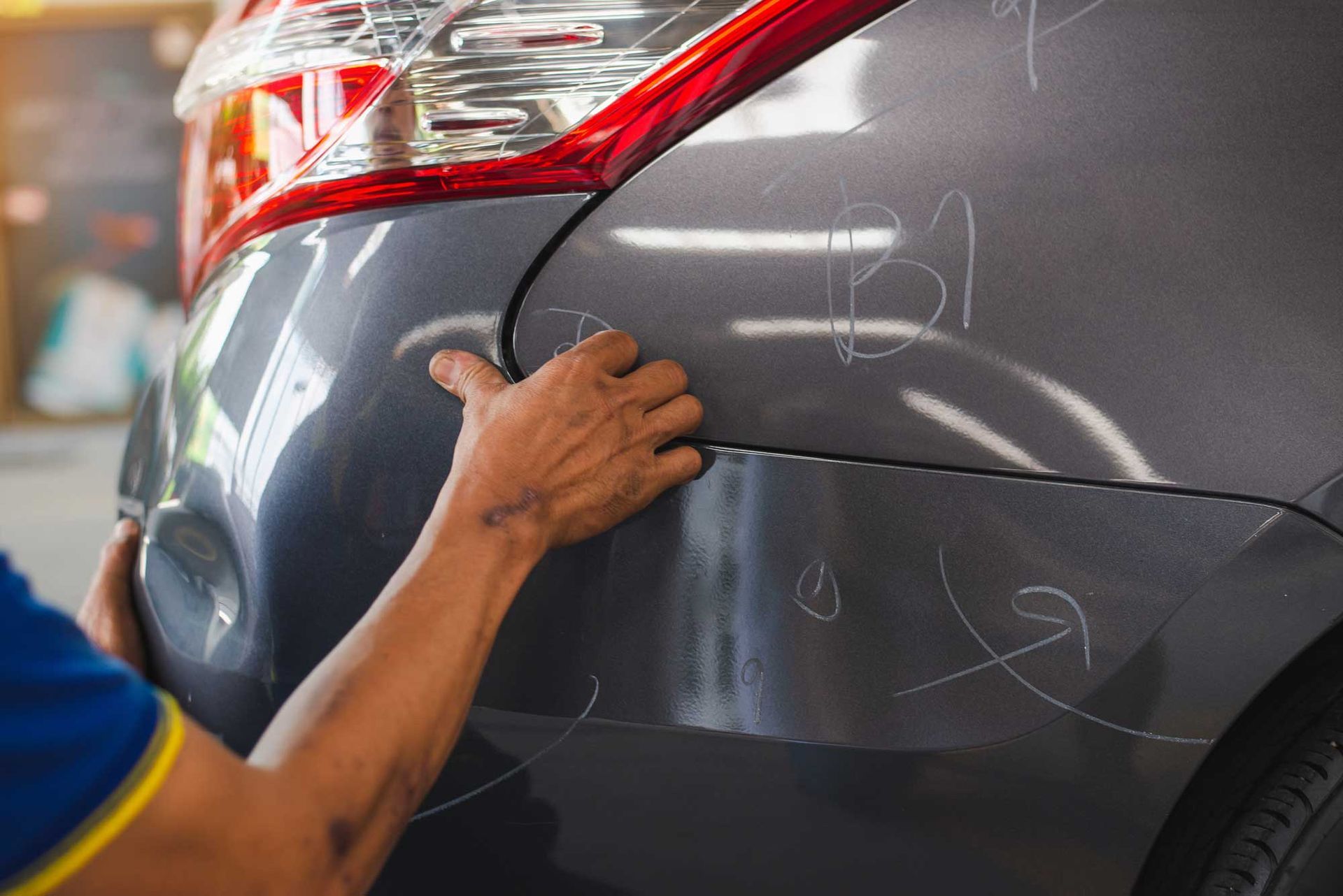 Person assessing car damage on the rear bumper, with visible scratches and dents. The car is grey, and in a repair shop.