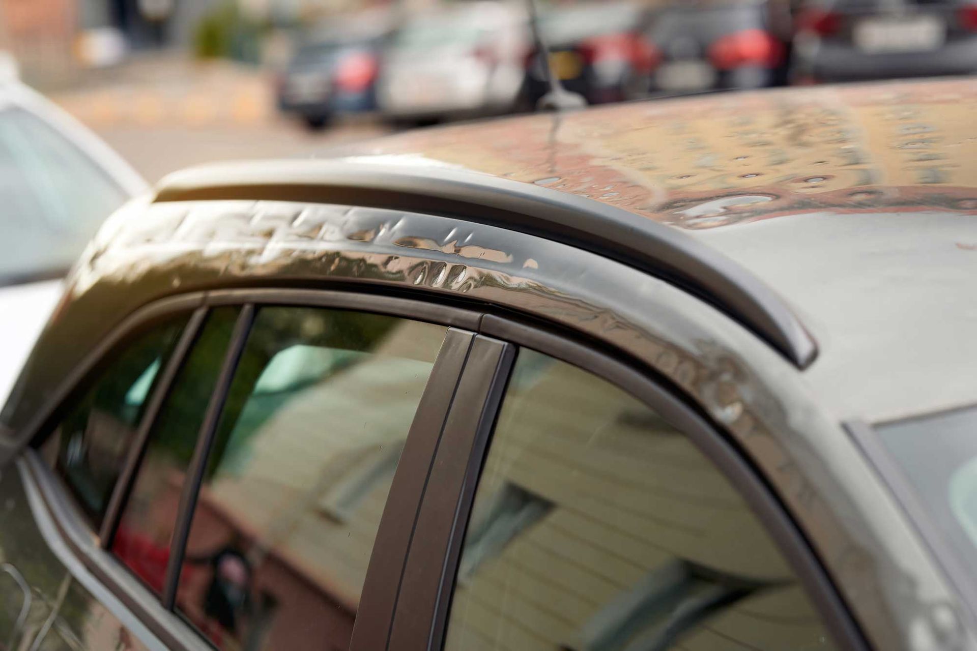 Close-up of a dark grey car roof with black roof rails and a side window, with blurry parked cars in the background.