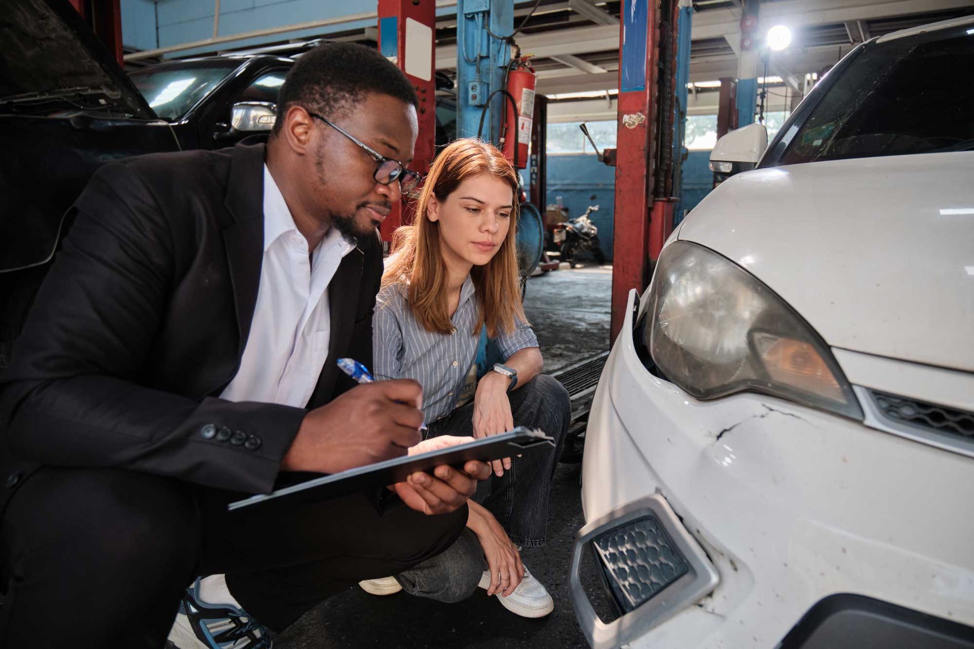 A mechanic in a suit inspects car damage with a customer, writing on a clipboard in a repair shop.