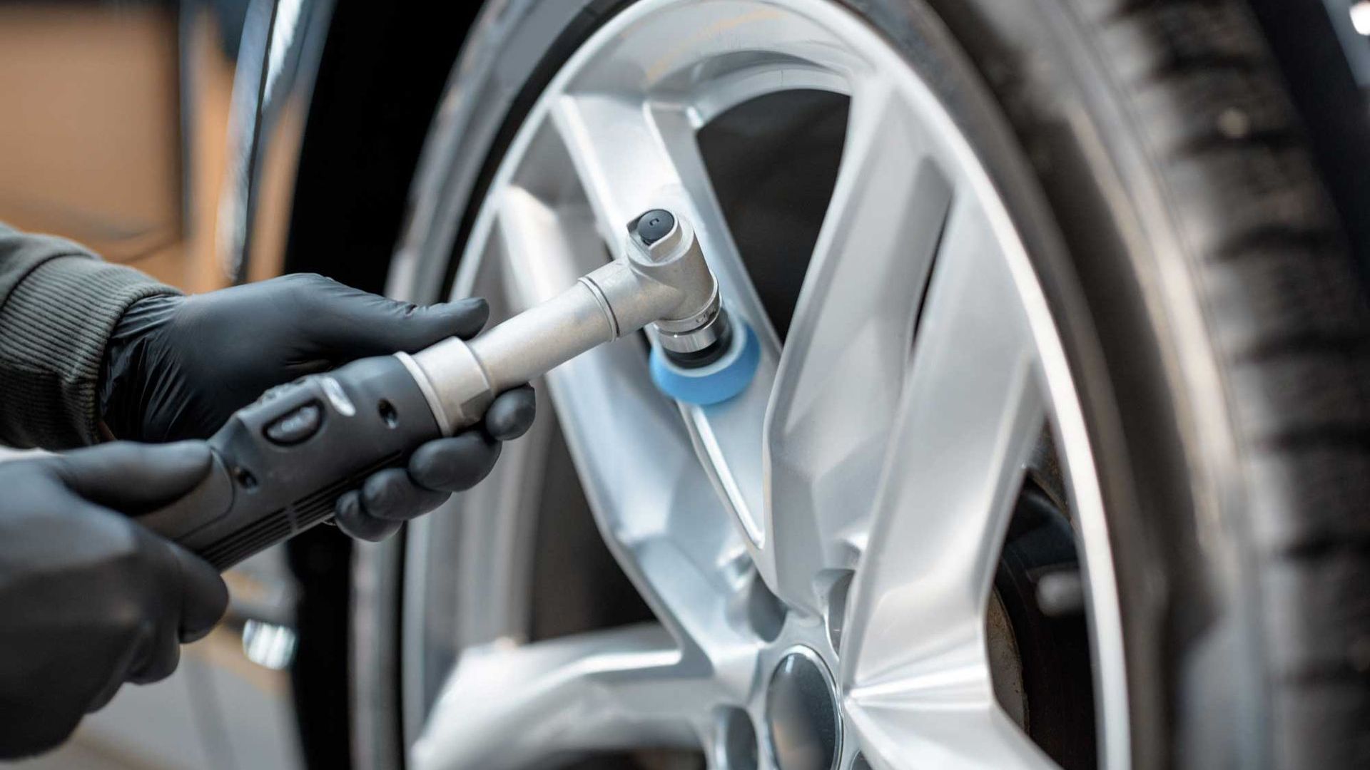 A person wearing black gloves polishes a silver car wheel with a handheld tool.