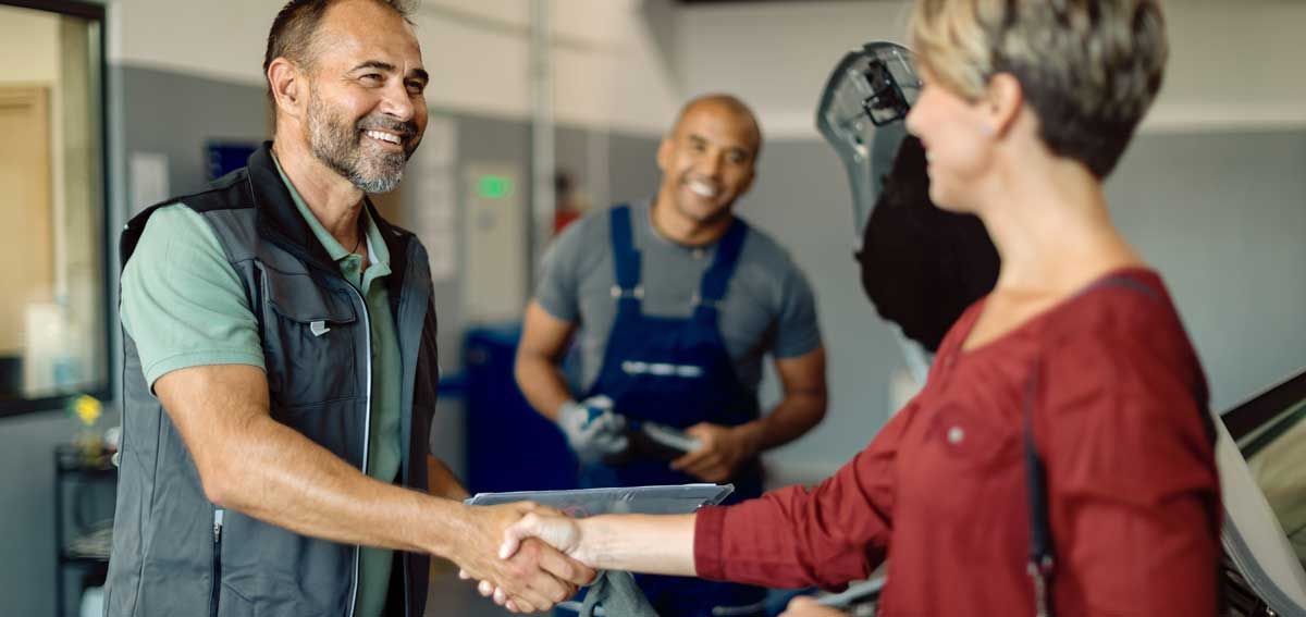 Two mechanics shake hands with a woman at a car repair shop; both smile. Another mechanic is in the background.