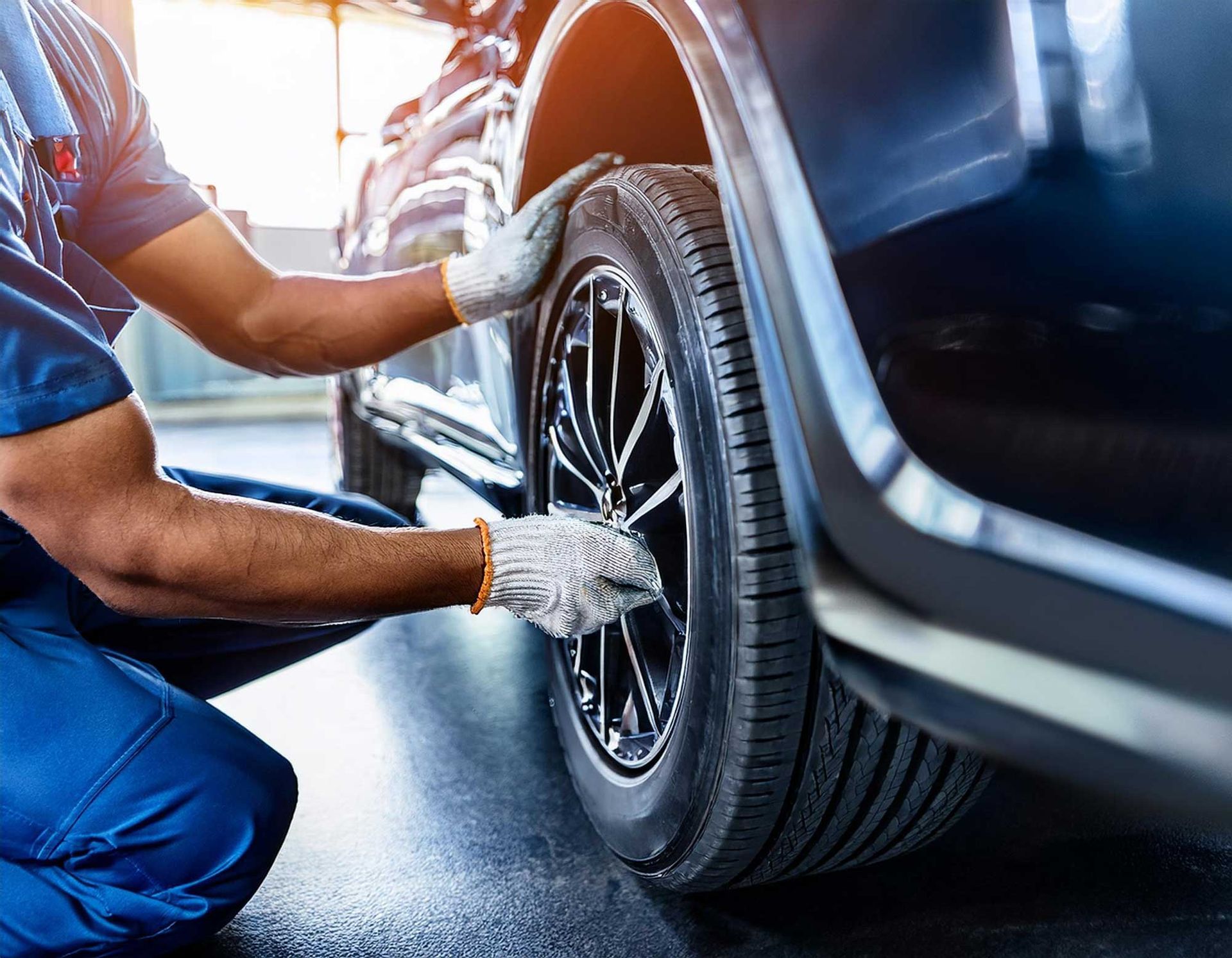 Mechanic inspecting a car tire, wearing gloves and blue work clothes, in a garage setting.