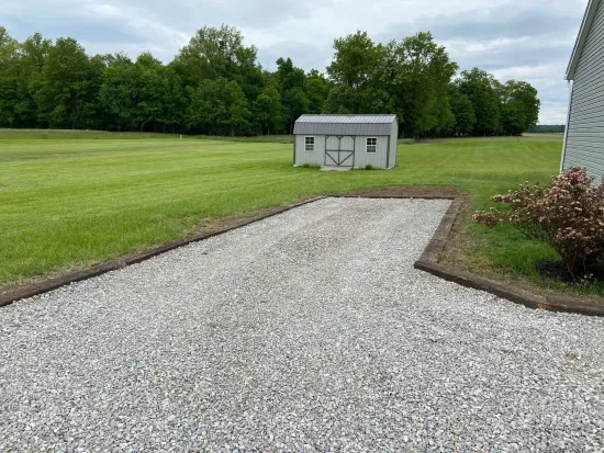 Gravel driveway bordered by wood leads to a small shed on a grassy yard. Trees in background.